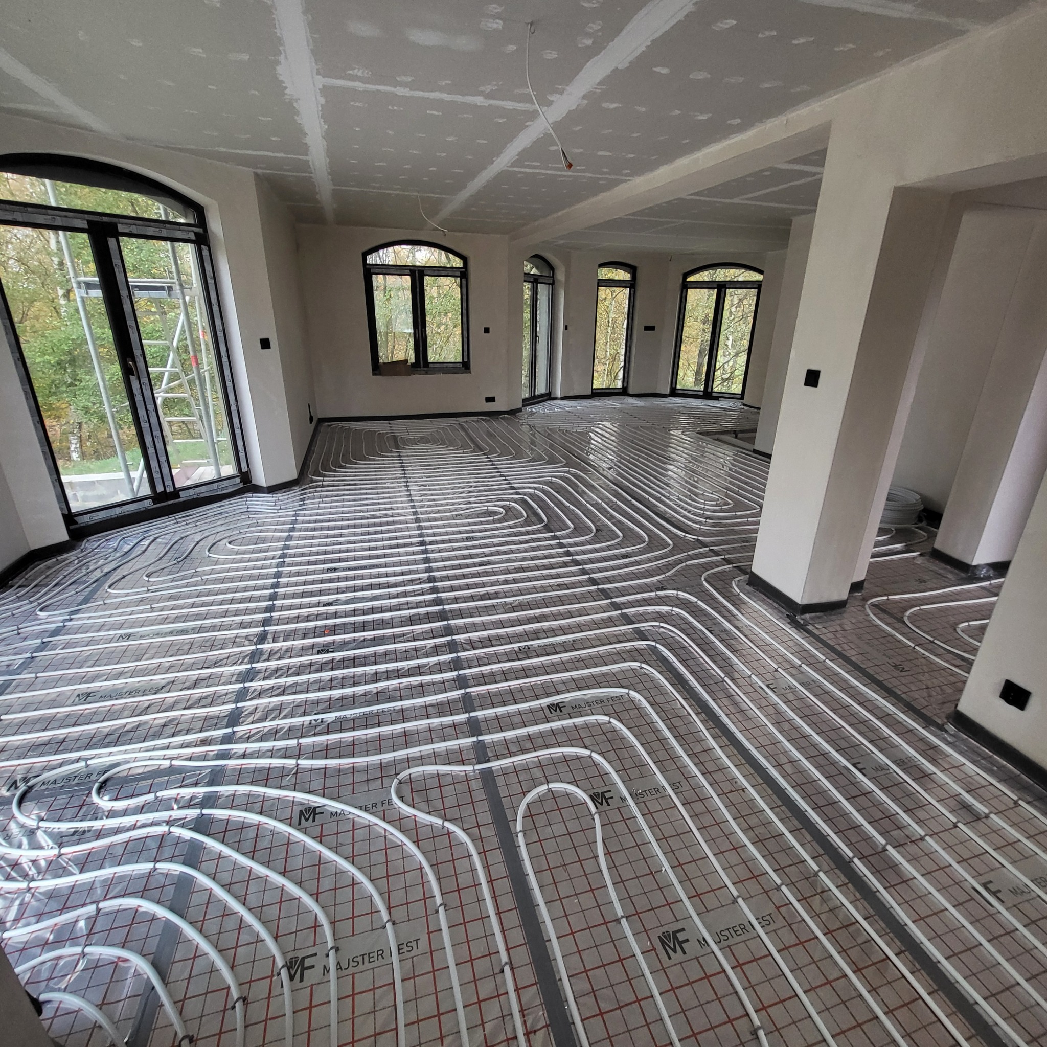 Białe rurki ogrzewania podłogowego ułożone na folii z nadrukiem w nowym, przestronnym pomieszczeniu z dużymi oknami i surowym wykończeniem ścian i sufitu.