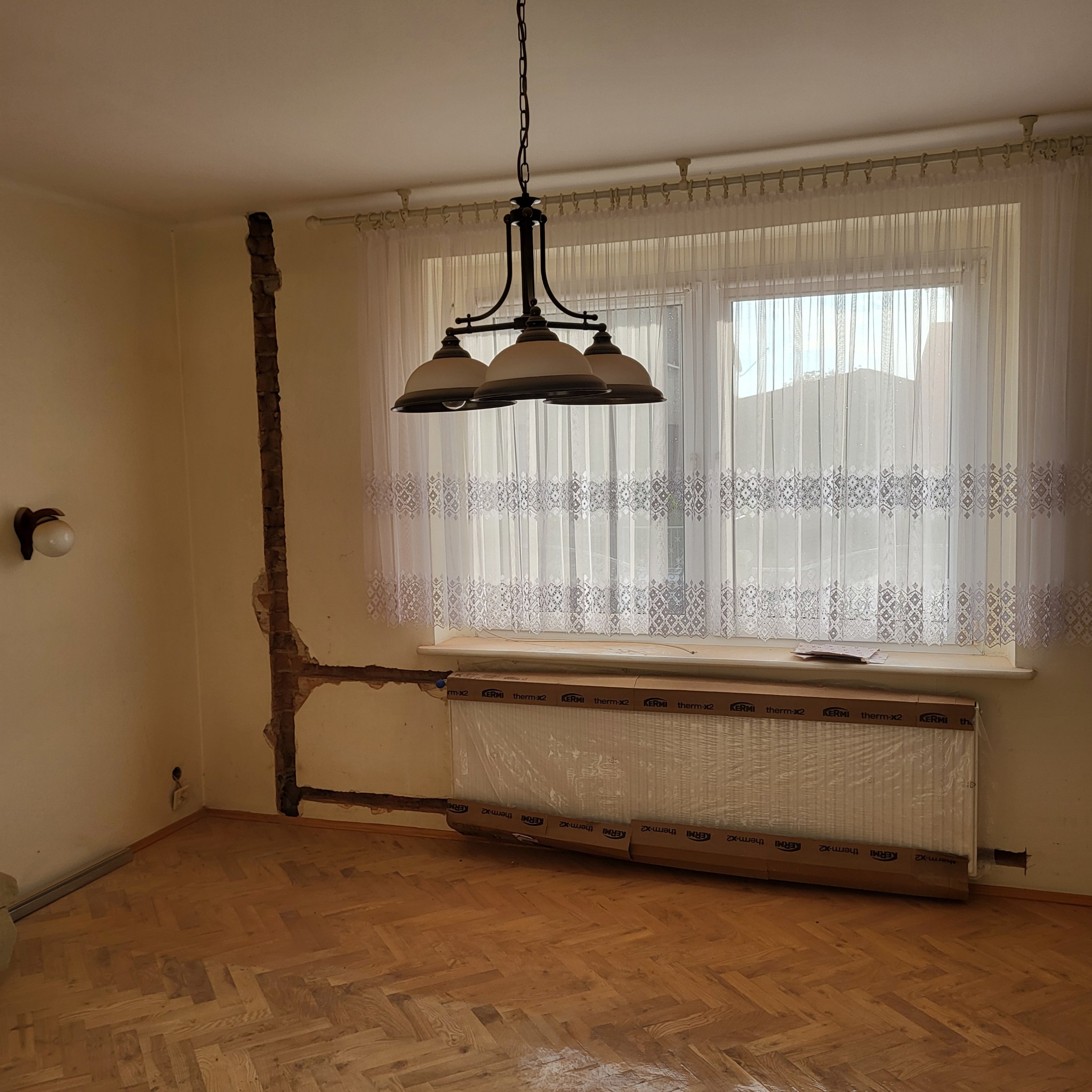 Pokój przed remontem instalacji grzewczej: odsłonięte fragmenty muru pod rury, nowy grzejnik w folii ochronnej marki KERMI, wisząca lampa z trzema kloszami, drewniany parkiet w jodełkę.