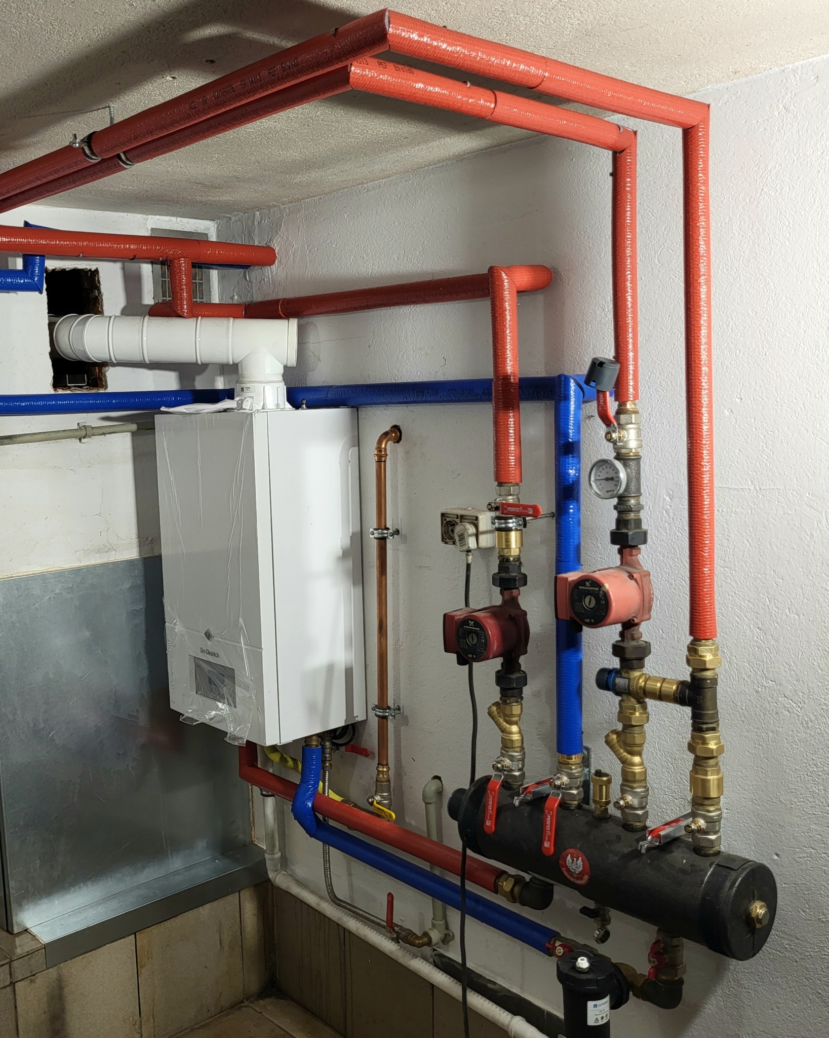 Nowy piec gazowy De Dietrich, jeszcze w folii ochronnej, podłączony do instalacji centralnego ogrzewania z czerwonymi izolowanymi rurami, niebieskimi rurami PCV i elementami pomiarowymi.