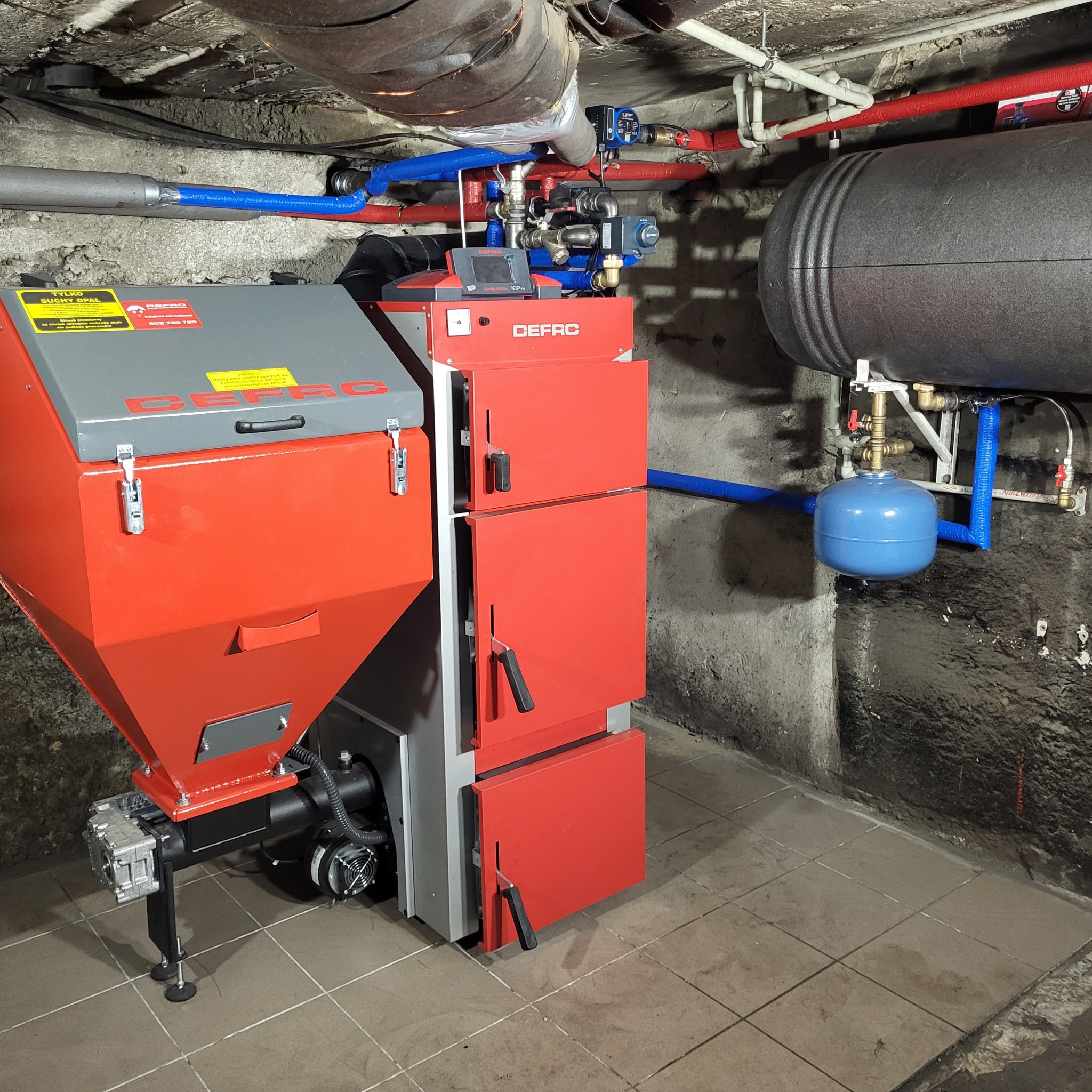 Czerwono-szary piec na paliwo stałe marki DEFRO z podajnikiem, zbiornikiem i sterownikiem, zainstalowany w piwnicy z widocznymi rurami instalacji grzewczej i zbiornikiem akumulacyjnym.