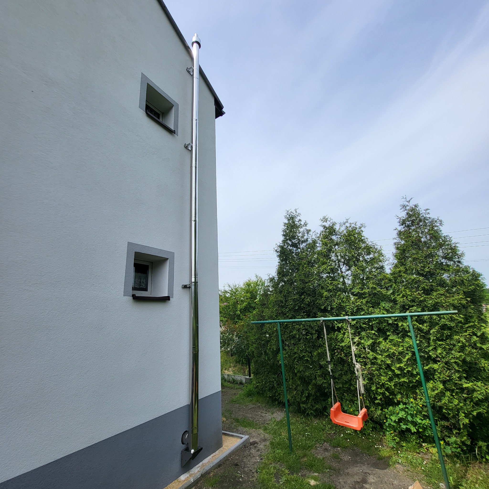 Zewnętrzny, pionowy, stalowy komin spalinowy przymocowany do ściany domu z widocznymi uchwytami mocującymi, na tle błękitnego nieba i zieleni przydomowego ogrodu z huśtawką.