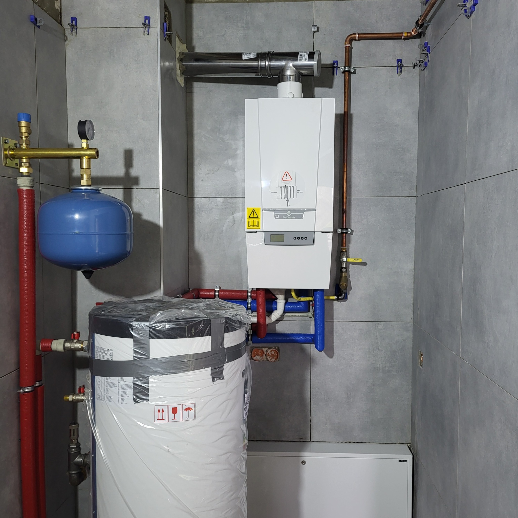 Nowoczesny piec gazowy De Dietrich z pionowym zasobnikiem ciepłej wody, rurami miedzianymi i niebieskim naczyniem przeponowym, zamontowany na szarej, płytkowej ścianie łazienki z widocznymi...