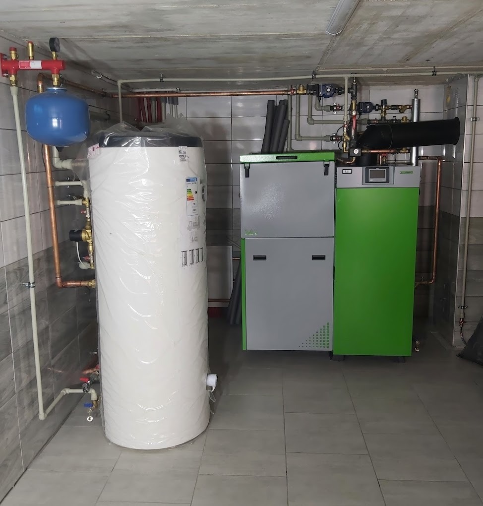 Kotłownia z piecem na paliwo stałe (eko-groszek) w kolorze szaro-zielonym oraz dużym, owiniętym folią ochronną, białym zasobnikiem ciepłej wody użytkowej. Instalacja z rur miedzianych i białych rur...