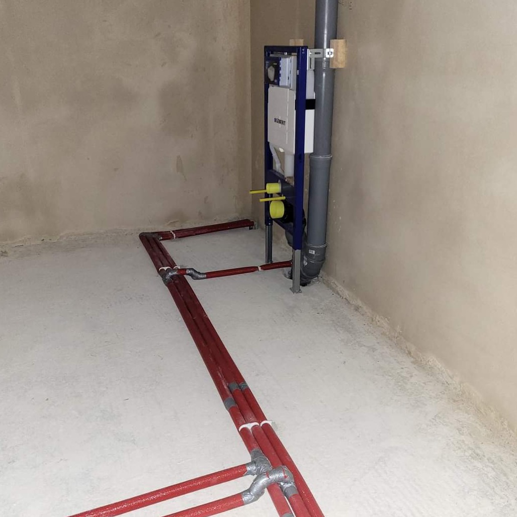 Surowy stan łazienki z widoczną instalacją wodną i kanalizacyjną przed montażem sanitariatów, rury w izolacji termicznej prowadzone po podłodze, stelaż podtynkowy Geberit zamocowany do ściany.