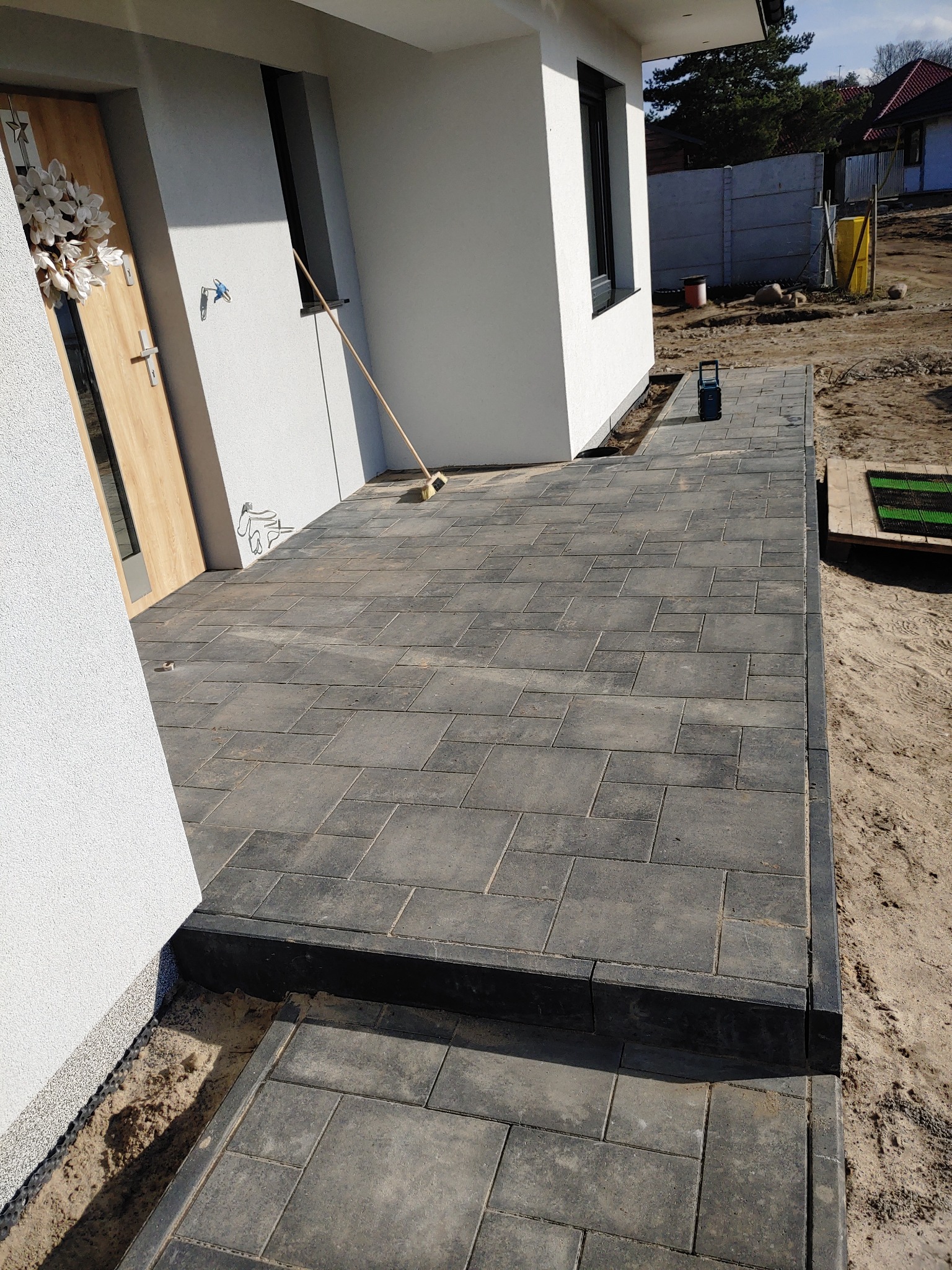 Nowo ułożony chodnik z szarej kostki brukowej prowadzący do wejścia domu z drewnianymi drzwiami ozdobionymi białym wieńcem, widoczna szczotka oparta o ścianę i laserowy niwelator na kostce.