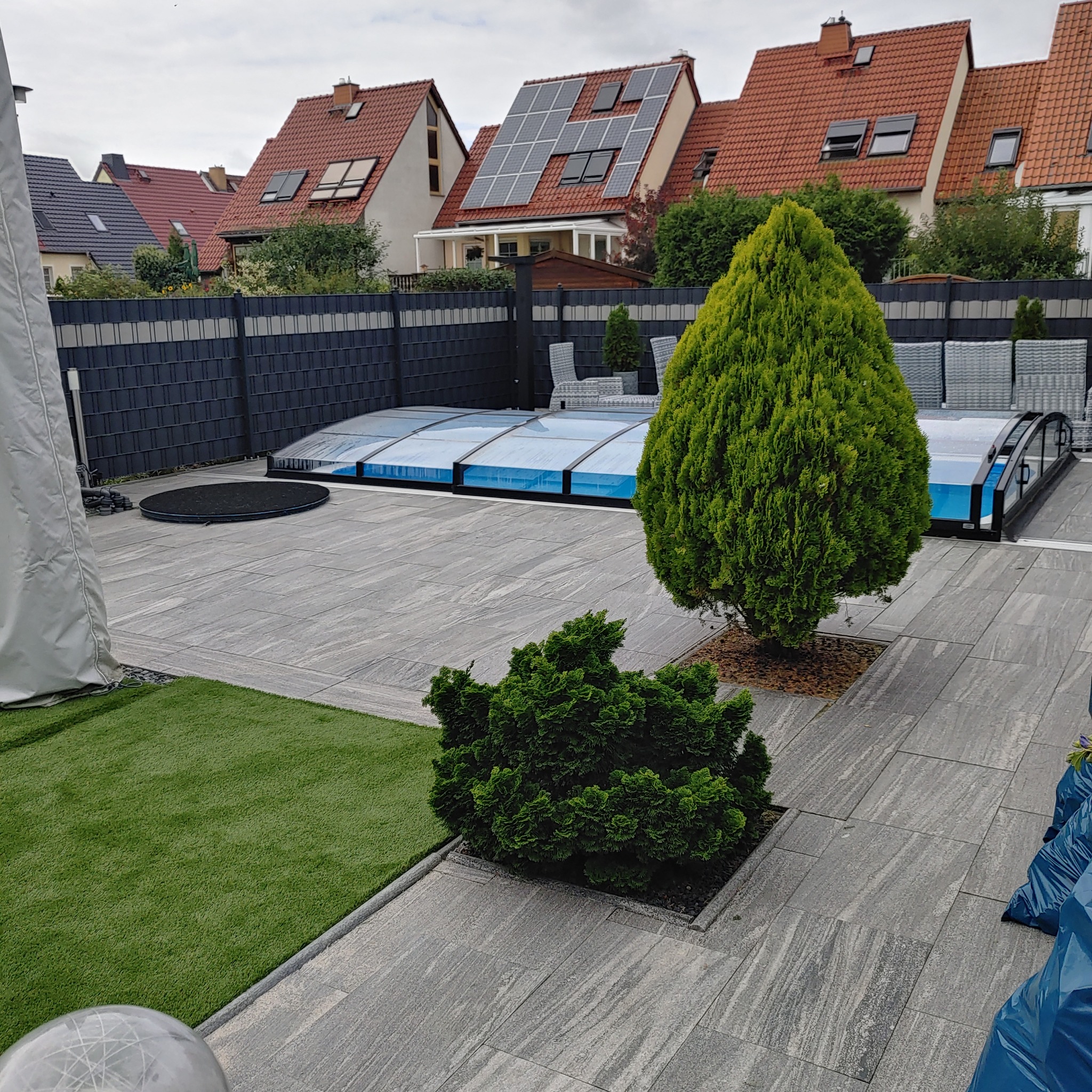 Szare, prostokątne płyty ułożone na tarasie z widocznym zadaszeniem basenu, tujami w kwadratowych donicach i sztuczną trawą, w tle domy jednorodzinne z panelami solarnymi na dachu.