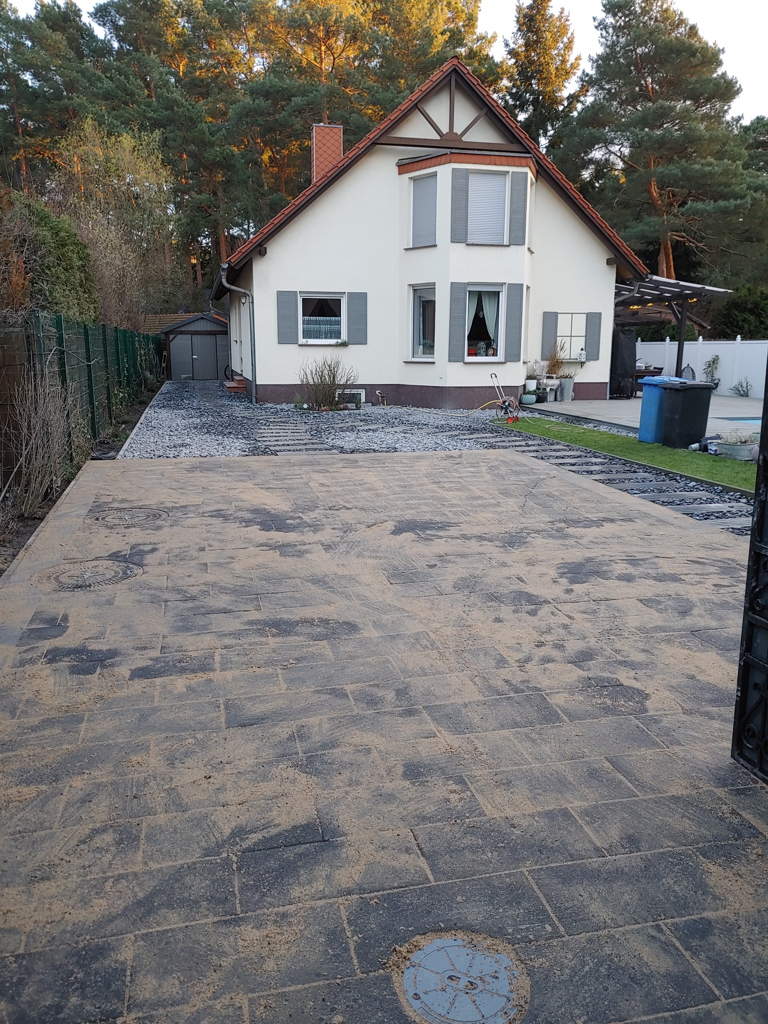 Nowa, duża powierzchnia z szarej kostki brukowej przed domem jednorodzinnym z białymi ścianami i szarymi okiennicami, widoczne studzienki kanalizacyjne, częściowo zasypana piaskiem, z boku ścieżka...