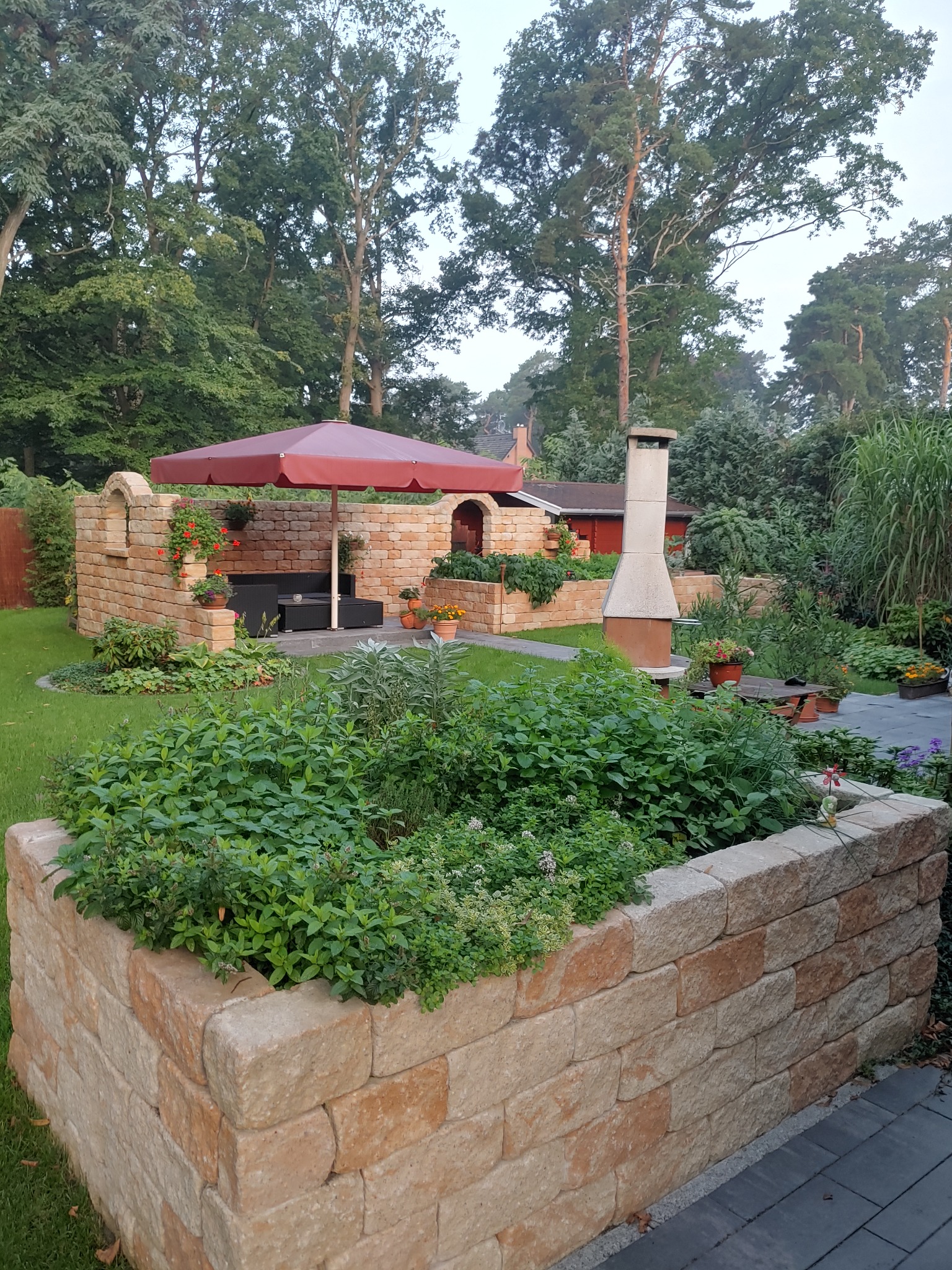 Ogród z podwyższonymi rabatami z piaskowca, zadaszonym miejscem do siedzenia i grillem kominkowym, otoczony zielenią.