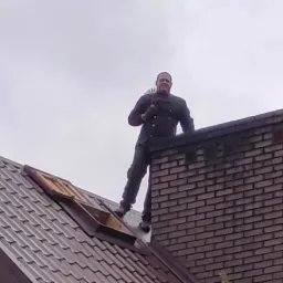 Uśmiechnięty mężczyzna w kombinezonie na dachu, stojący obok ceglanego komina z narzędziem w ręku, widok z dołu na tle pochmurnego nieba.