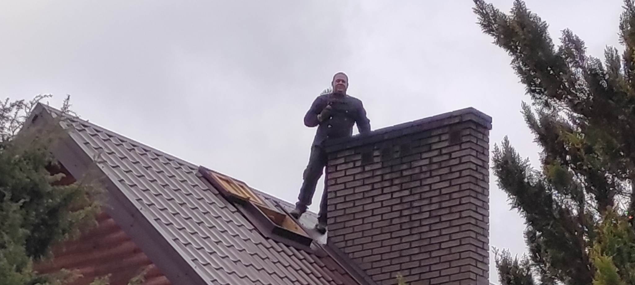Uśmiechnięty mężczyzna w kombinezonie na dachu, stojący obok ceglanego komina z narzędziem w ręku, widok z dołu na tle pochmurnego nieba.