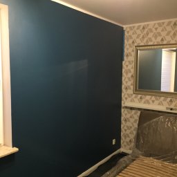 ArtDesign - Pokój po malowaniu: ściana w kolorze głębokiego granatu, zabezpieczone folią meble, fragment ściany z geometryczną tapetą i lustrem w złotej ramie.