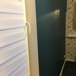 ArtDesign - Świeżo pomalowana, ciemnoturkusowa ściana z widocznym załamaniem światła, sąsiadująca z oknem z białymi żaluzjami i fragmentem ściany z geometryczną tapetą.