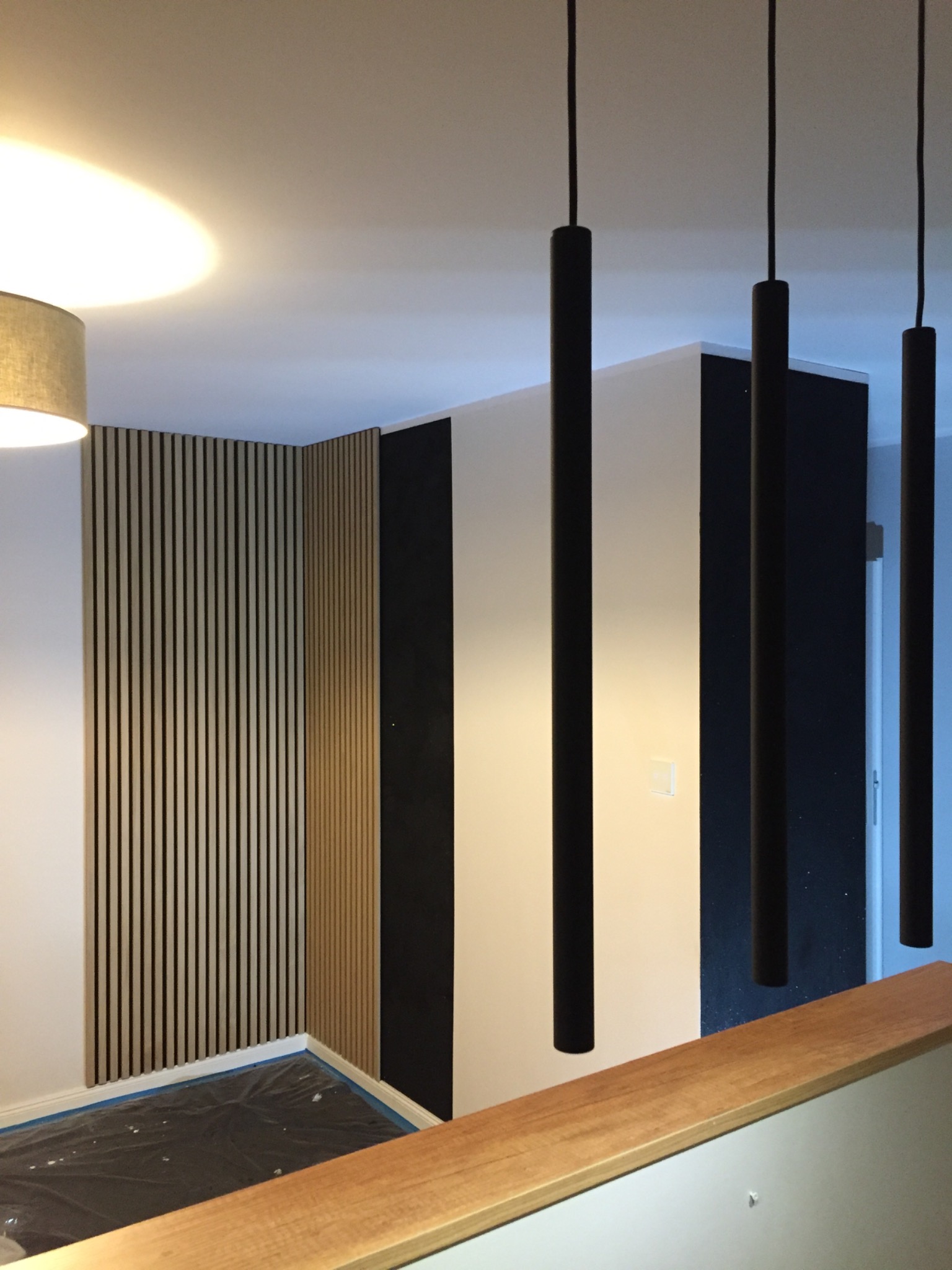 Nowoczesne wykończenie wnętrza z drewnianą boazerią lamelową na ścianie, minimalistyczne czarne lampy wiszące i fragment drewnianej balustrady, podłoga zabezpieczona folią malarską.