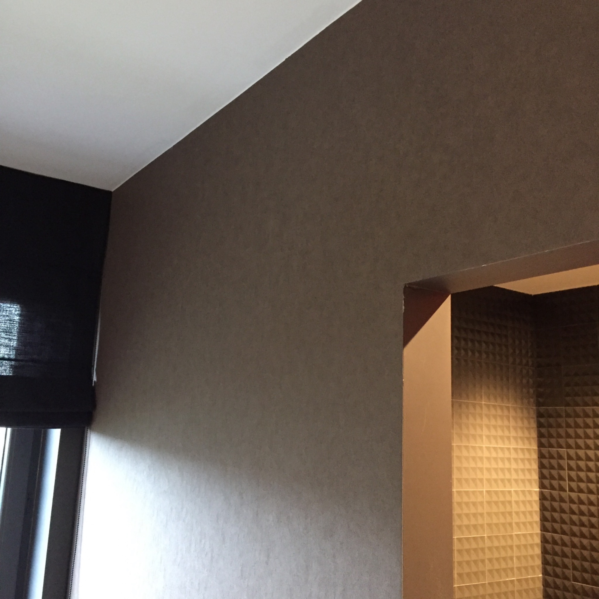 Ściana pokryta tapetą w odcieniach brązu, widoczna roleta okienna i fragment łazienki z geometrycznymi płytkami, w tle światło