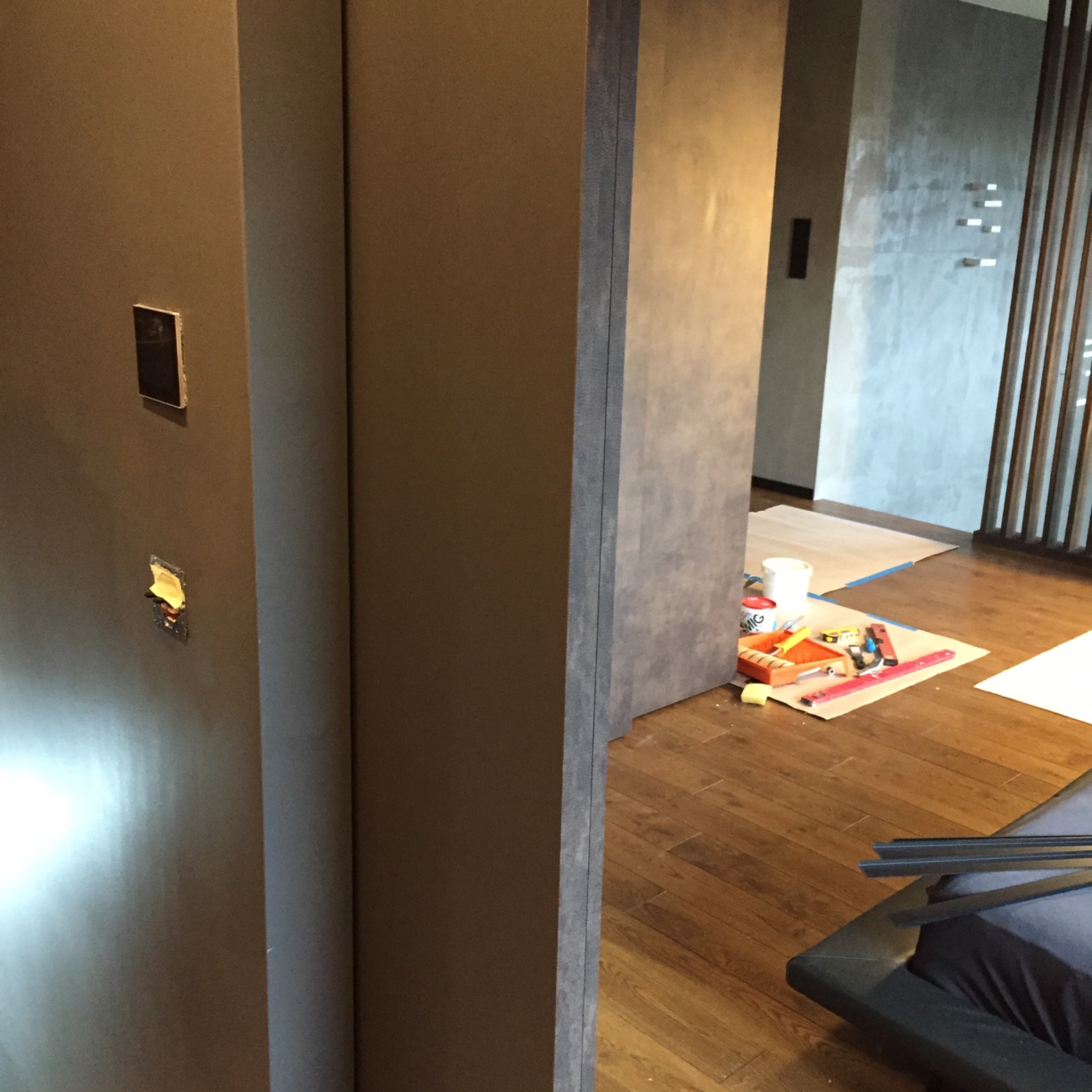 Wnętrze w trakcie malowania: widoczne narzędzia malarskie na zabezpieczonej podłodze, przygotowana ściana z niedokończonym gniazdkiem elektrycznym, surowa szara ściana w tle.