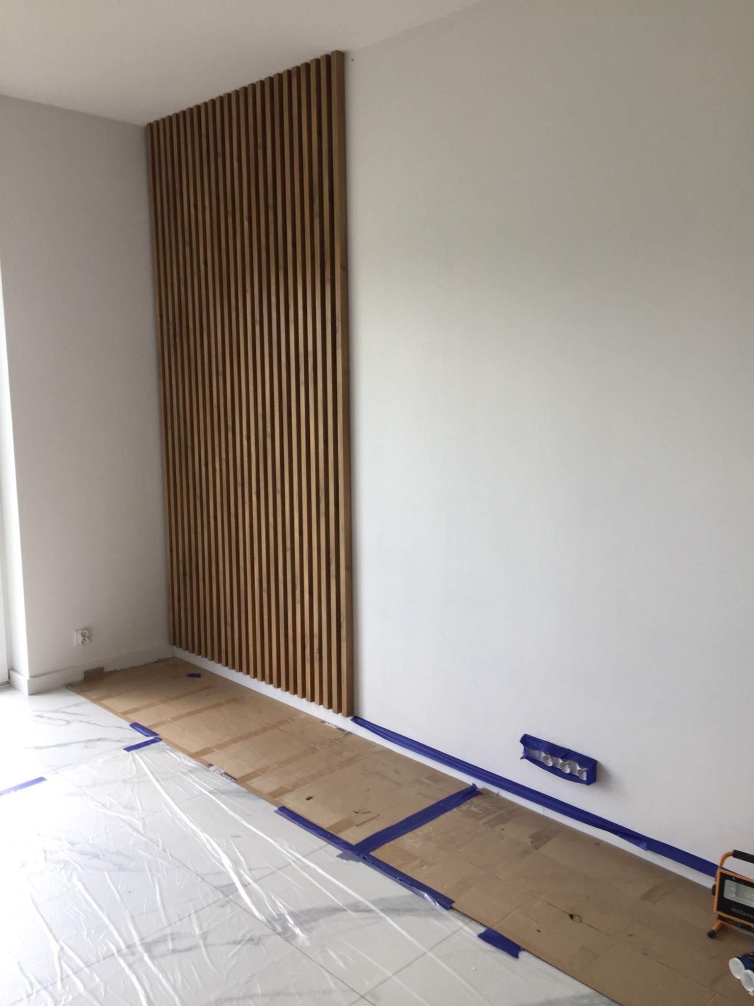 Montaż dekoracyjnych, pionowych paneli drewnianych na ścianie w trakcie wykańczania wnętrza, zabezpieczona podłoga folią i kartonem, widoczne gniazdko elektryczne i lampa budowlana.
