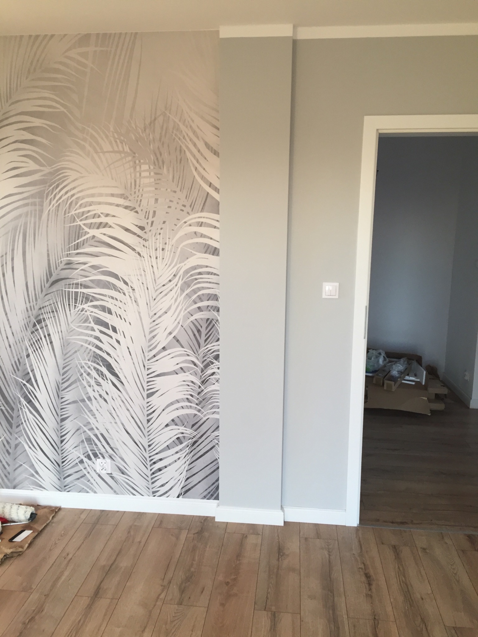 Ściana z szaro-białą tapetą w motywy roślinne, widoczny fragment podłogi z paneli, drzwi do innego pomieszczenia z kartonami w środku