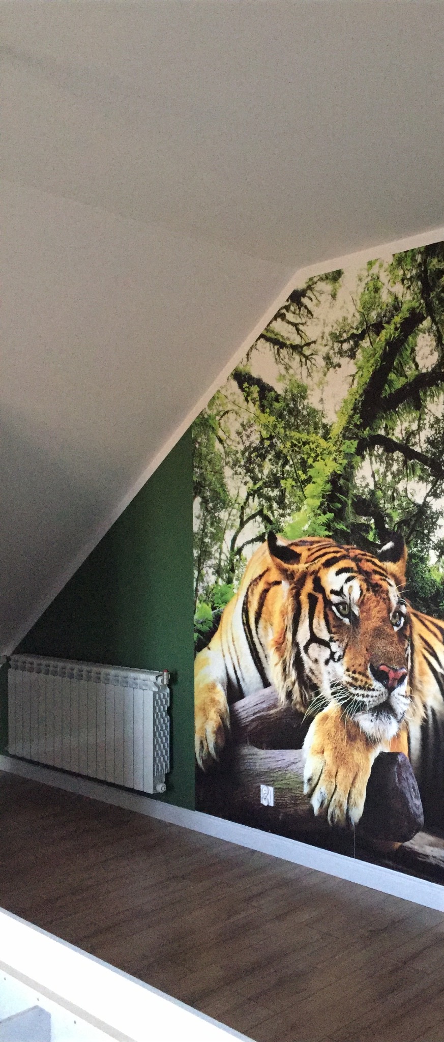 Pokój na poddaszu z fototapetą przedstawiającą tygrysa leżącego na pniu drzewa, zielona ściana, biały grzejnik i drewniana podłoga.