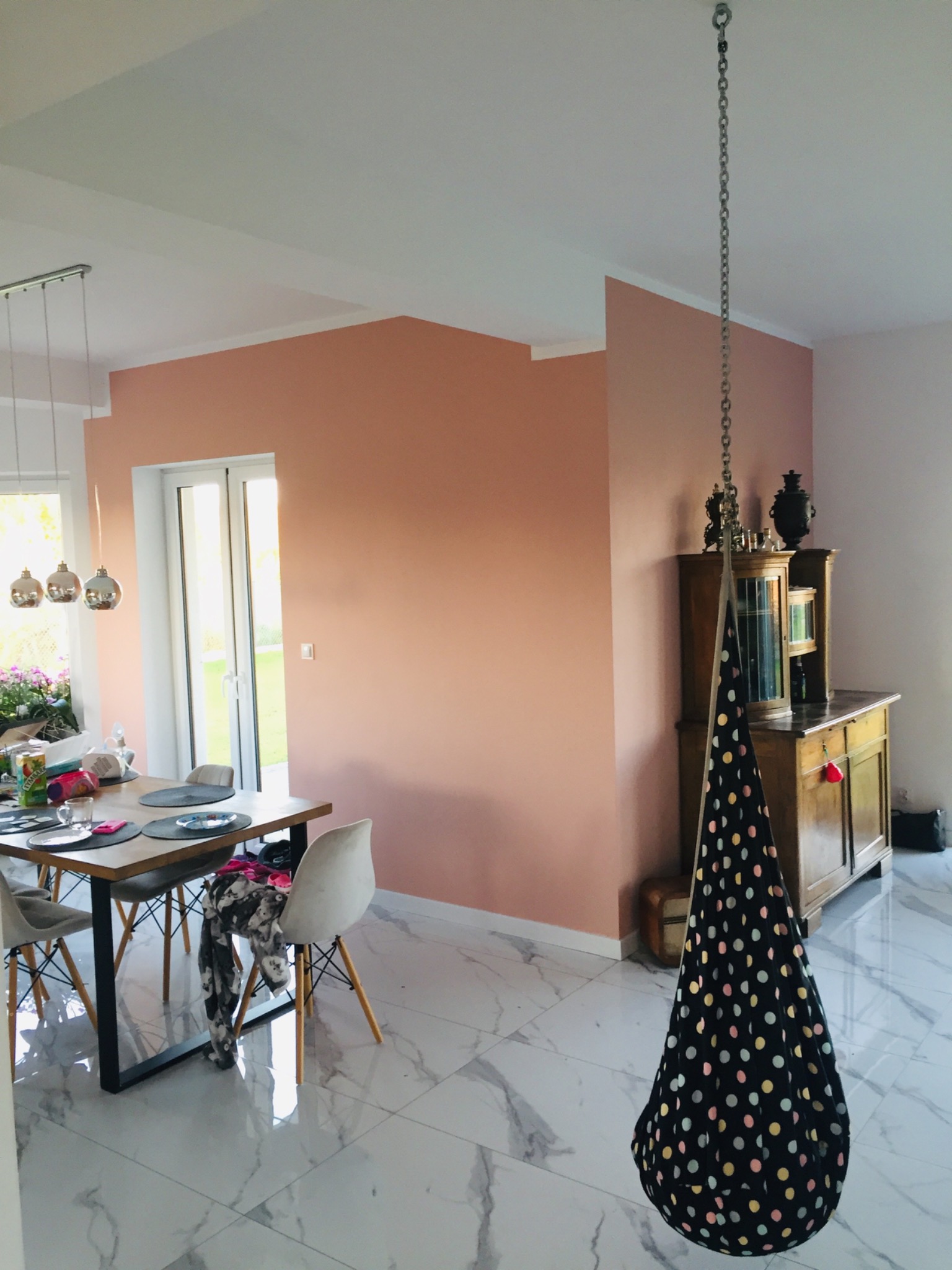 Jasne wnętrze z marmurową podłogą, ścianą pomalowaną na łososiowy kolor, drewnianą komodą z dekoracjami i zawieszonym na łańcuchu czarnym siedziskiem w kolorowe grochy.