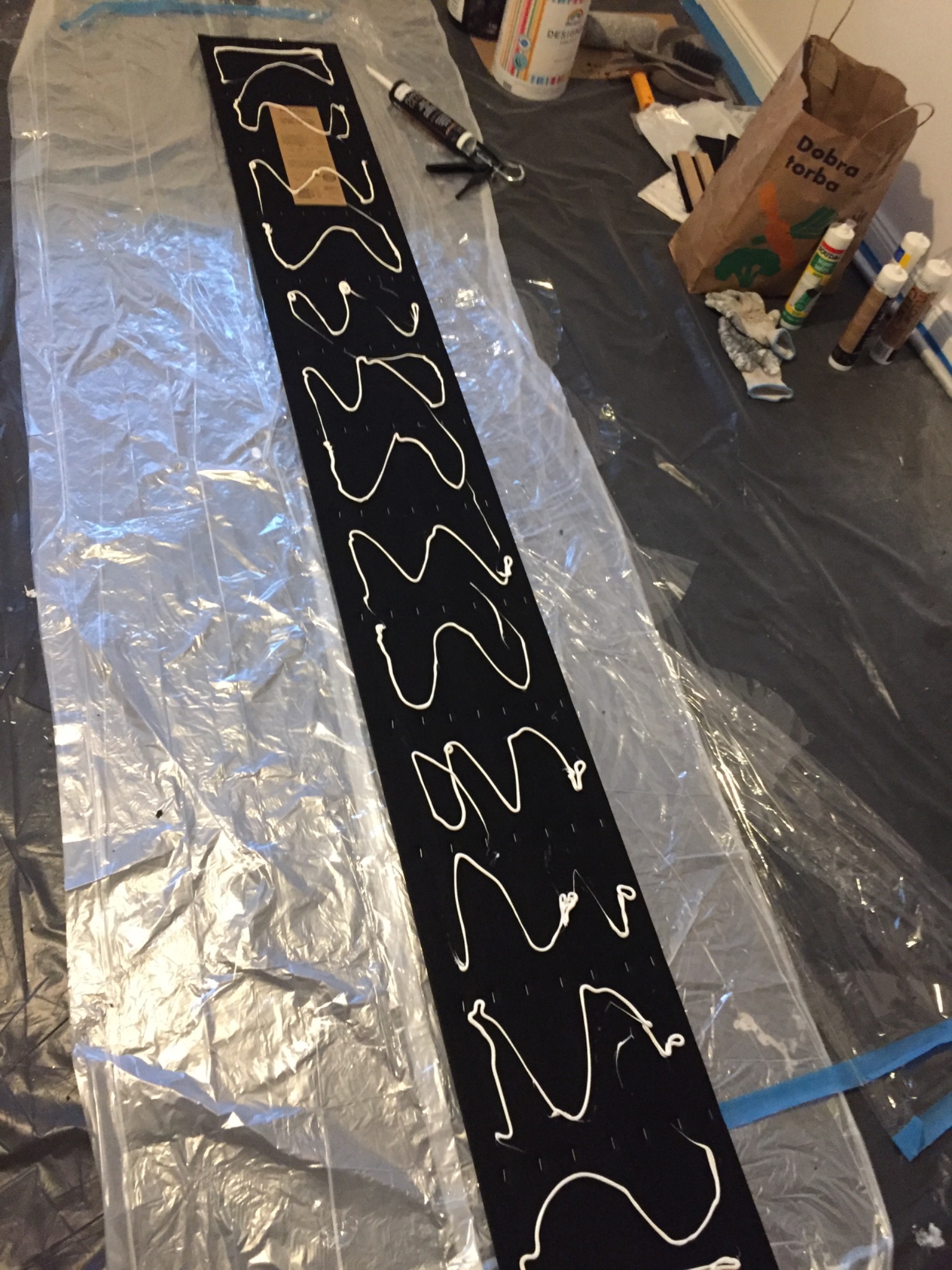 Czarny panel z nawierconymi otworami i nałożonym zygzakowatym wzorem z białego kleju, leżący na folii ochronnej, obok narzędzia do aplikacji kleju i torby z napisem 'Dobra torba'.