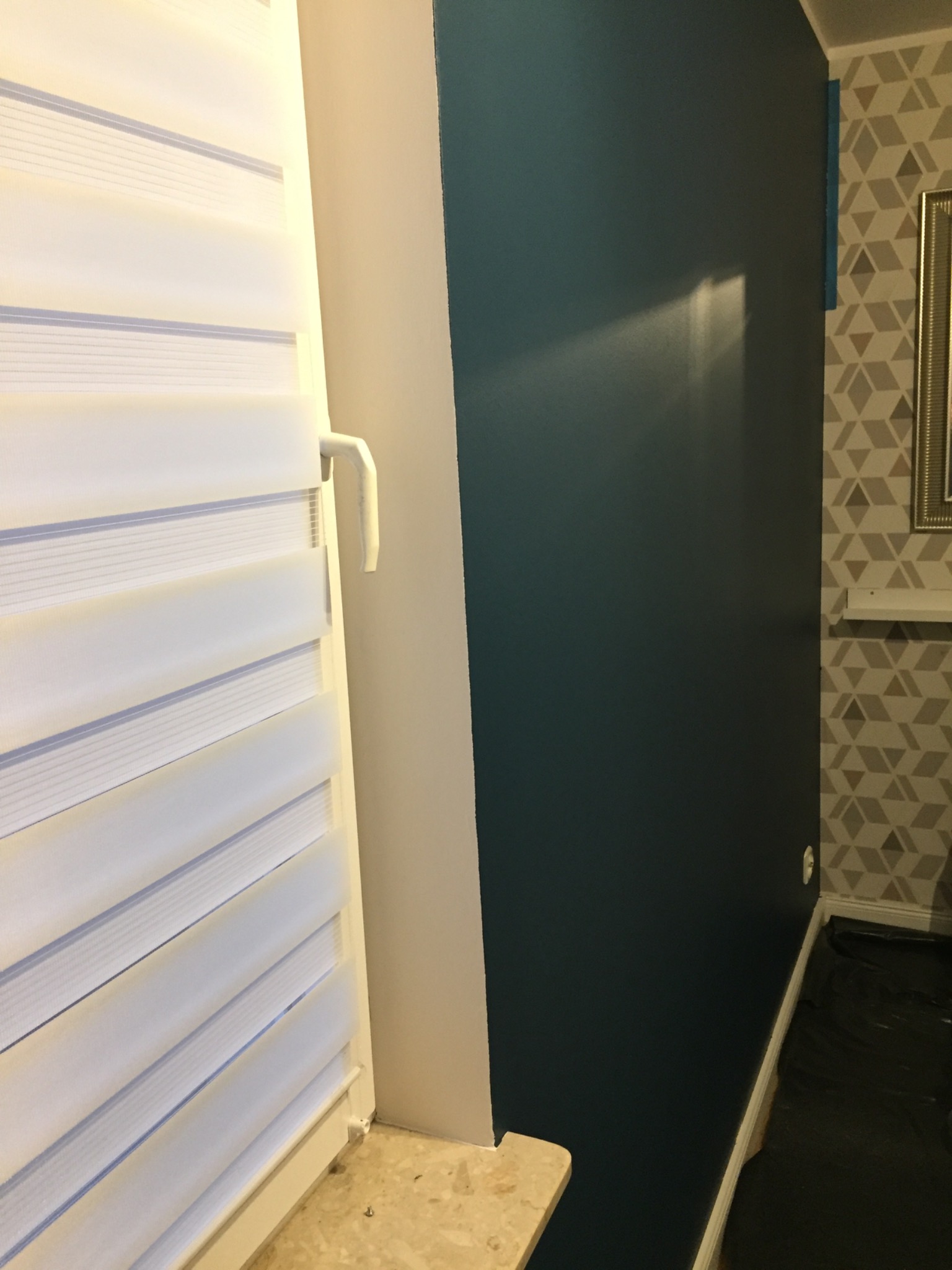 Świeżo pomalowana, ciemnoturkusowa ściana z widocznym załamaniem światła, sąsiadująca z oknem z białymi żaluzjami i fragmentem ściany z geometryczną tapetą.