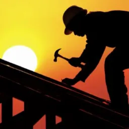 Sylwetka dekarza pracującego na więźbie dachowej na tle zachodzącego słońca, trzymającego młotek i wbijającego gwóźdź.