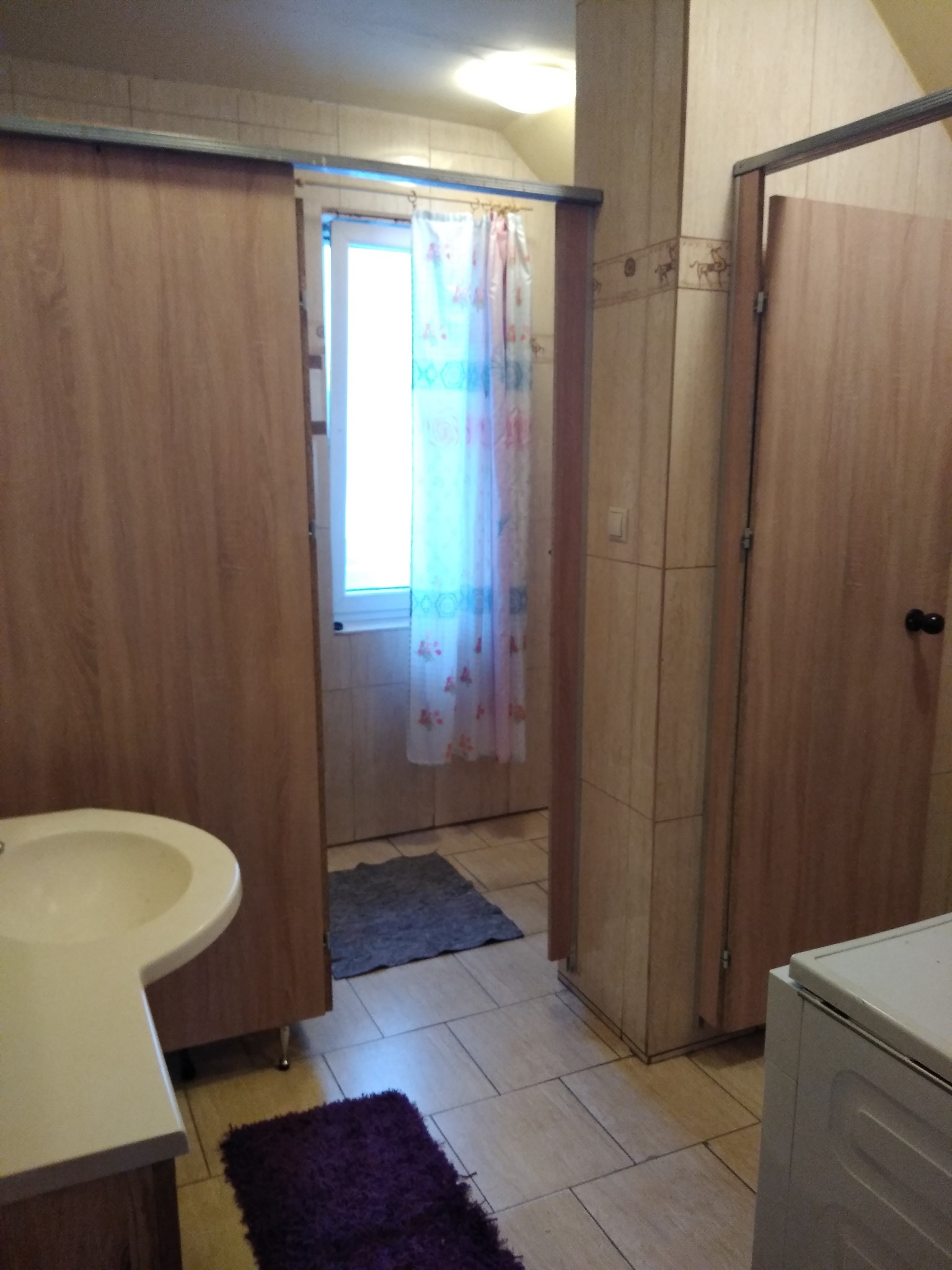 Wnętrze łazienki z umywalką, widok na okno z zasłoną i drzwi do kabiny prysznicowej, na podłodze dywaniki.