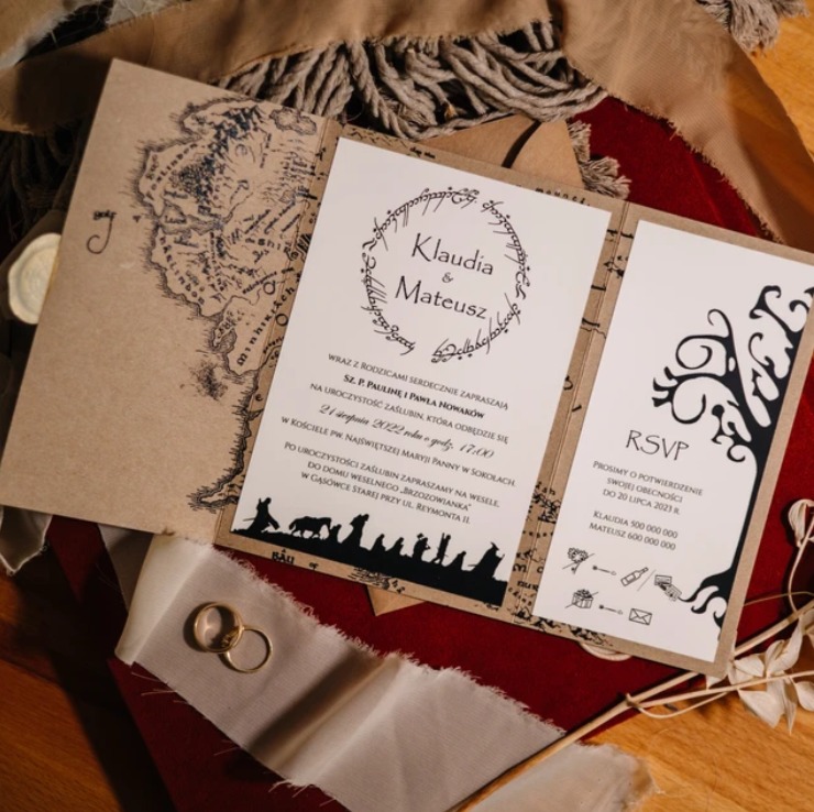 Otwarta, składana kartka zaproszenia ślubnego z motywami inspirowanymi Władcą Pierścieni, leżąca na czerwonej tkaninie i jutowym sznurku, obok para złotych obrączek