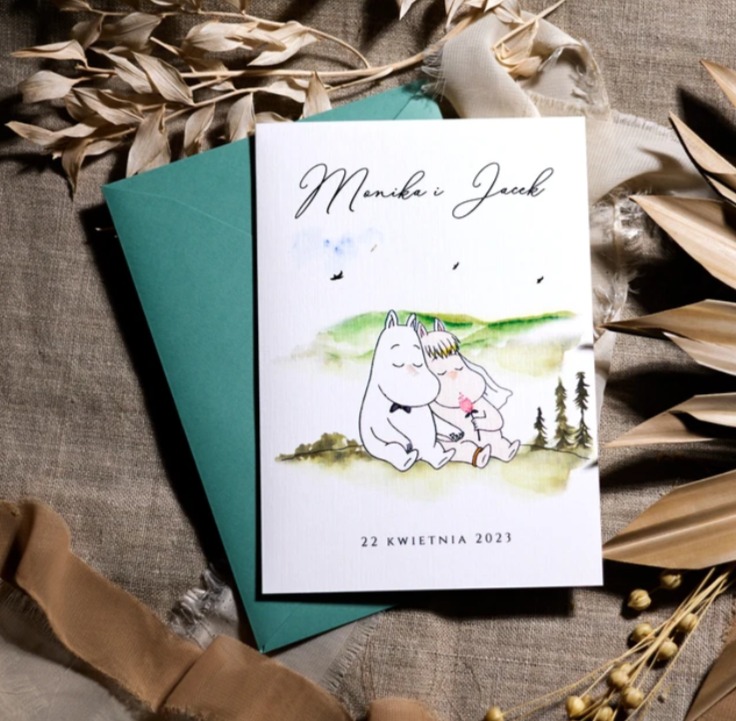 Zaproszenie ślubne z motywem Muminków dla Moniki i Jacka, datowane na 22 kwietnia 2023, w pastelowych kolorach, leżące na lnianym tle w Krakowie, obok zielonej koperty i suszonych roślin.