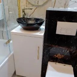 Elegancka łazienka z umywalką nablatową w kolorze czarnego marmuru, złotą armaturą i białymi meblami na tle marmurowych płytek.