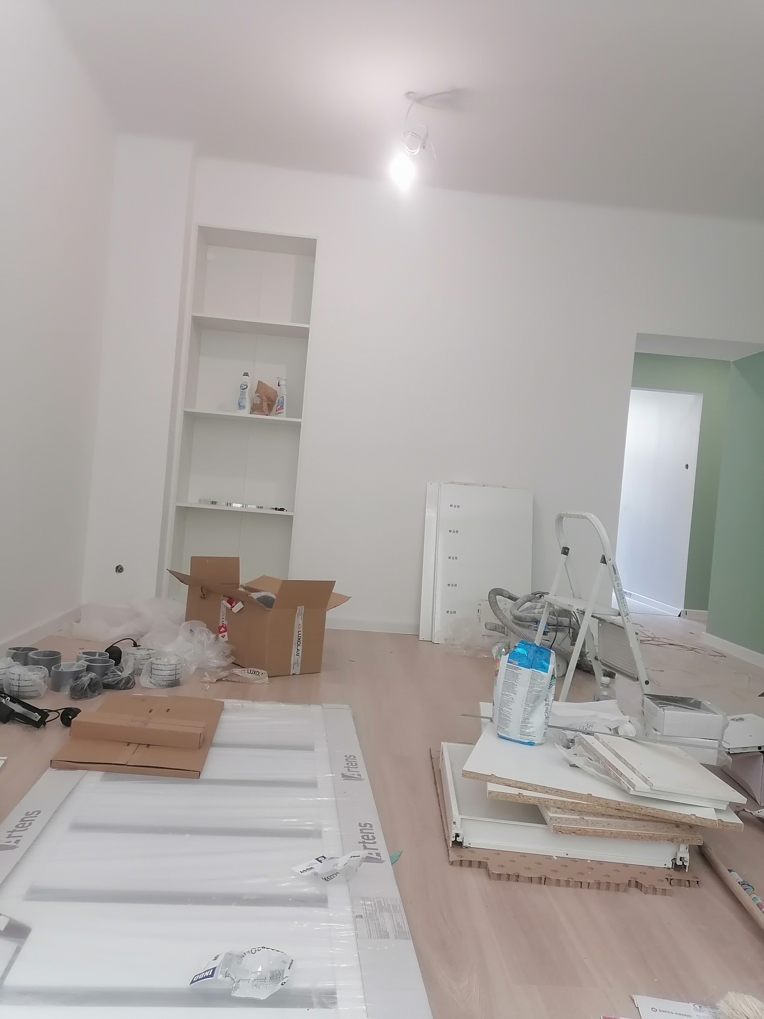 Wnętrze w trakcie prac wykończeniowych: białe ściany, regał wnękowy, rozpakowane elementy meblowe, drabina, kartony i akcesoria budowlane na podłodze.