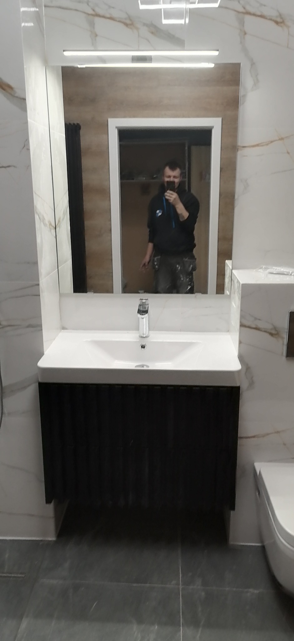 Umywalka z czarną szafką i lustrem z podświetleniem LED w wykończonej łazience w Łodzi, widoczne marmurowe płytki na ścianach i szare płytki podłogowe, odbicie fotografa w lustrze.