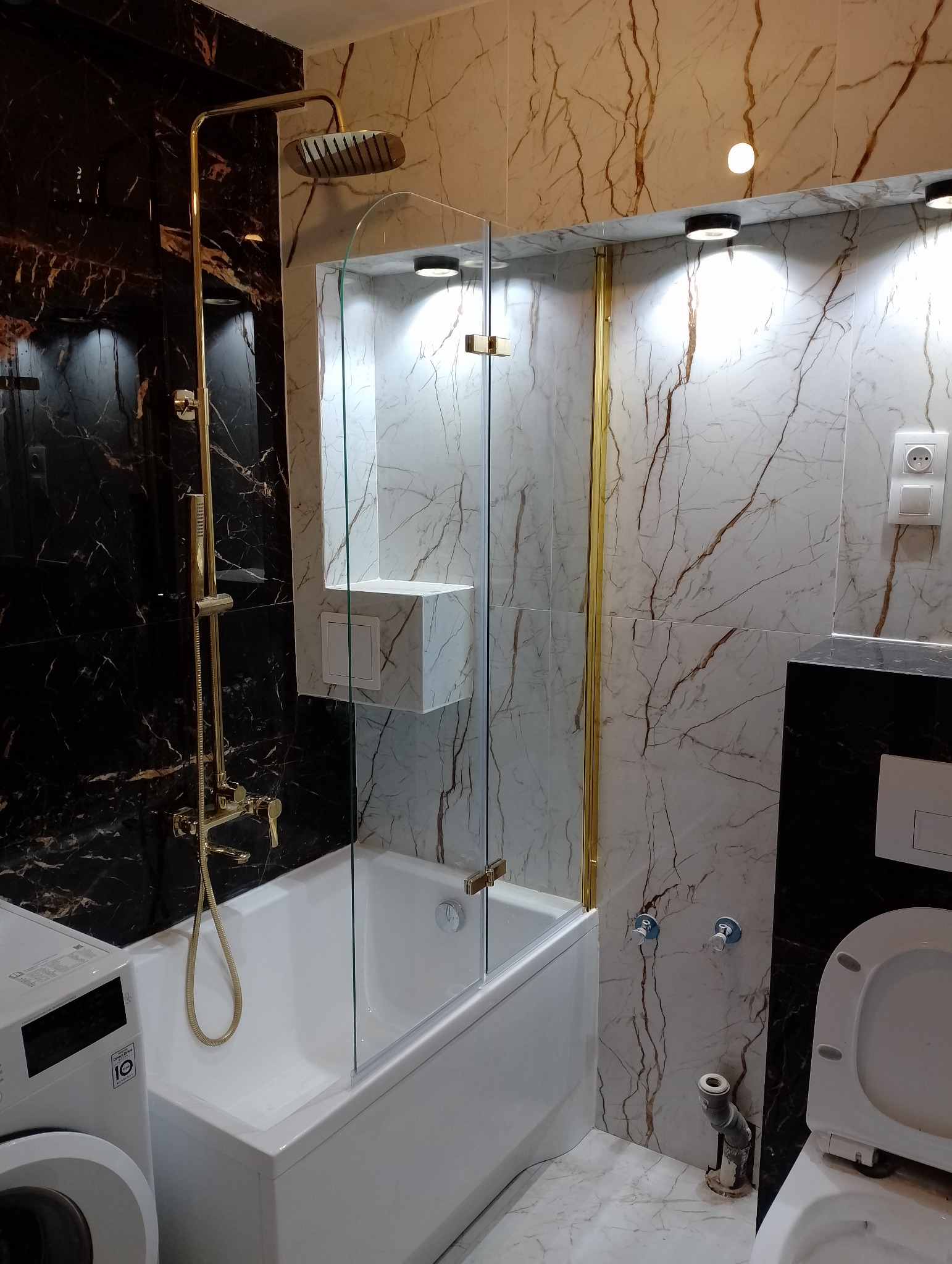 Nowoczesna łazienka z marmurowymi płytkami, złotą armaturą, kabiną prysznicową ze szklanymi drzwiami, pralką i toaletą.