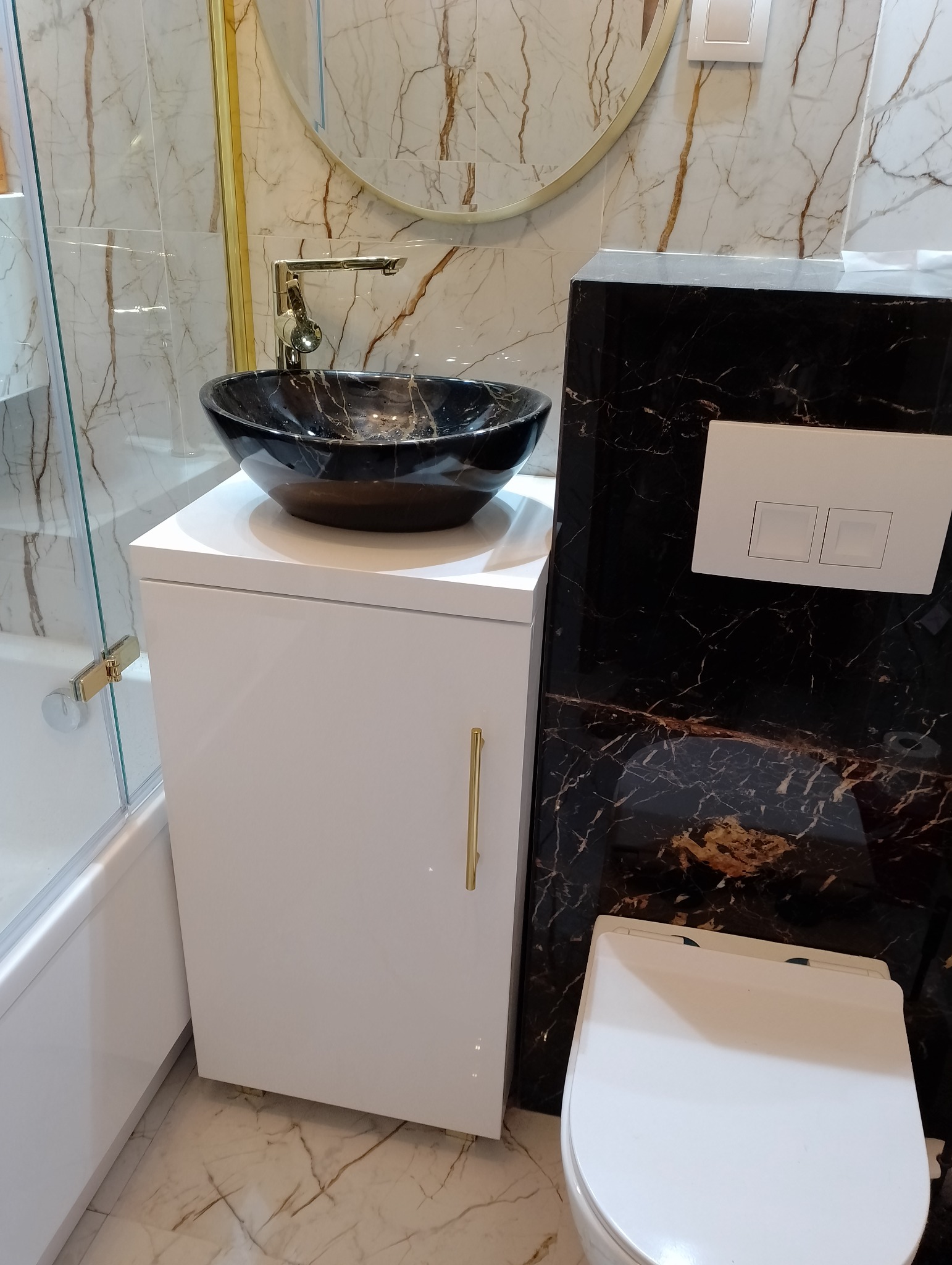 Elegancka łazienka z umywalką nablatową w kolorze czarnego marmuru, złotą armaturą i białymi meblami na tle marmurowych płytek.