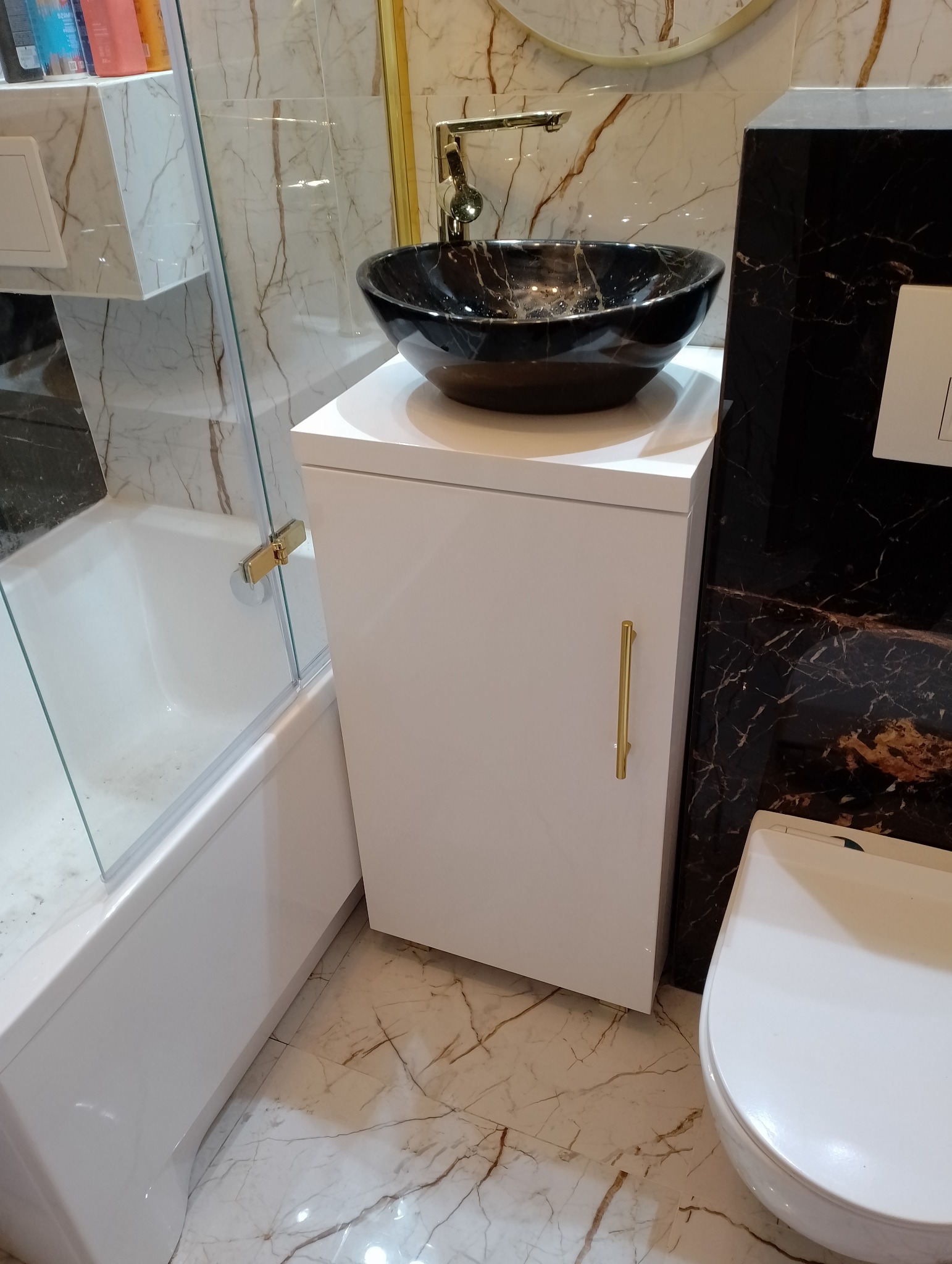 Nowoczesna łazienka z umywalką nablatową w kolorze czarnego marmuru, złotą armaturą i marmurowymi płytkami w odcieniach bieli i brązu, z kabiną prysznicową i białą szafką pod umywalką.