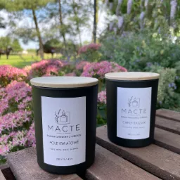 Dwie czarne świece sojowe z drewnianymi wieczkami na drewnianym stole, z kwiatami i zielenią w tle, jedna z etykietą 'Pole Kwiatowe', druga 'Ciepły Kaszmir'.