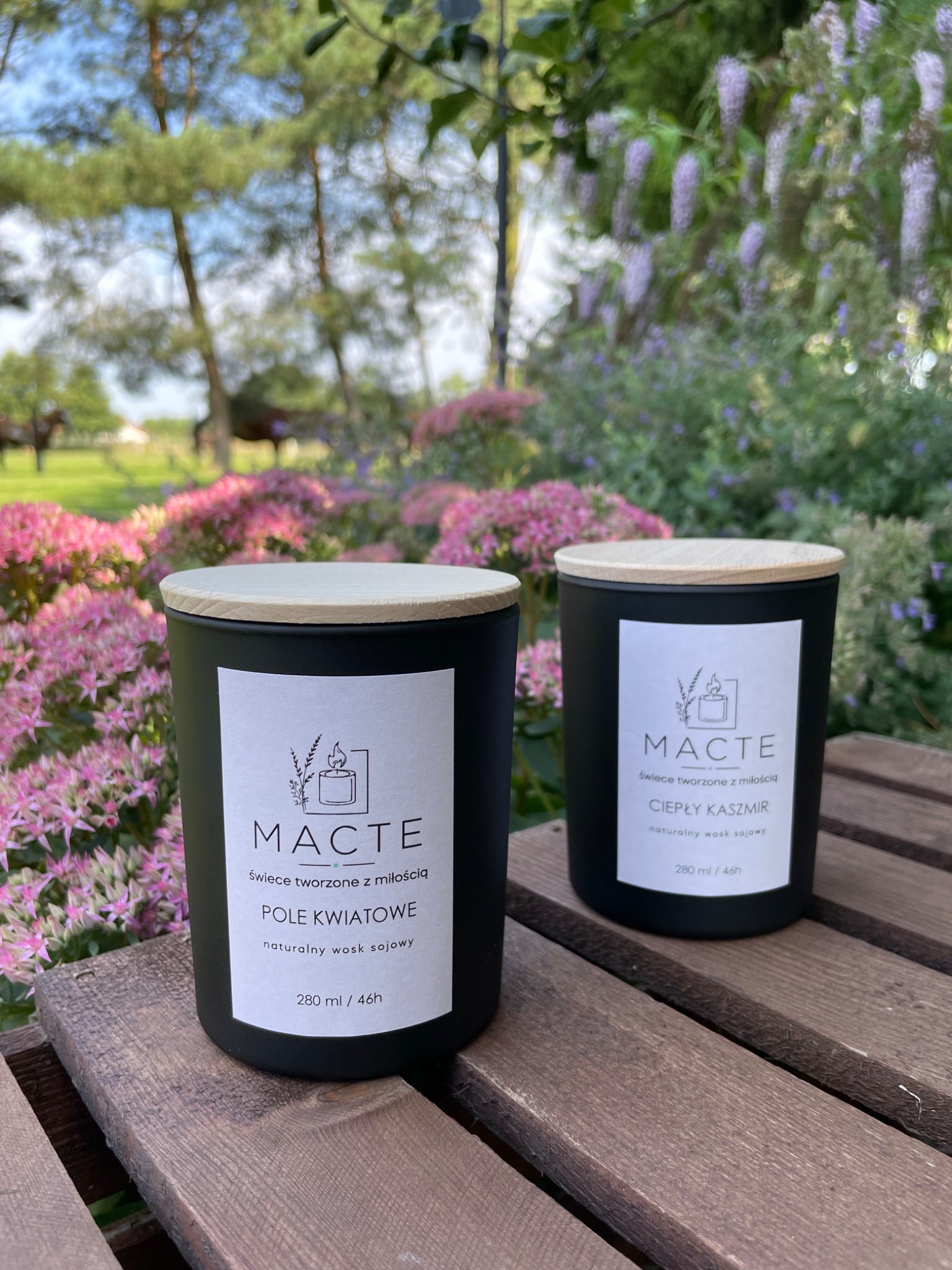 Dwie czarne świece sojowe z drewnianymi wieczkami na drewnianym stole, z kwiatami i zielenią w tle, jedna z etykietą 'Pole Kwiatowe', druga 'Ciepły Kaszmir'.