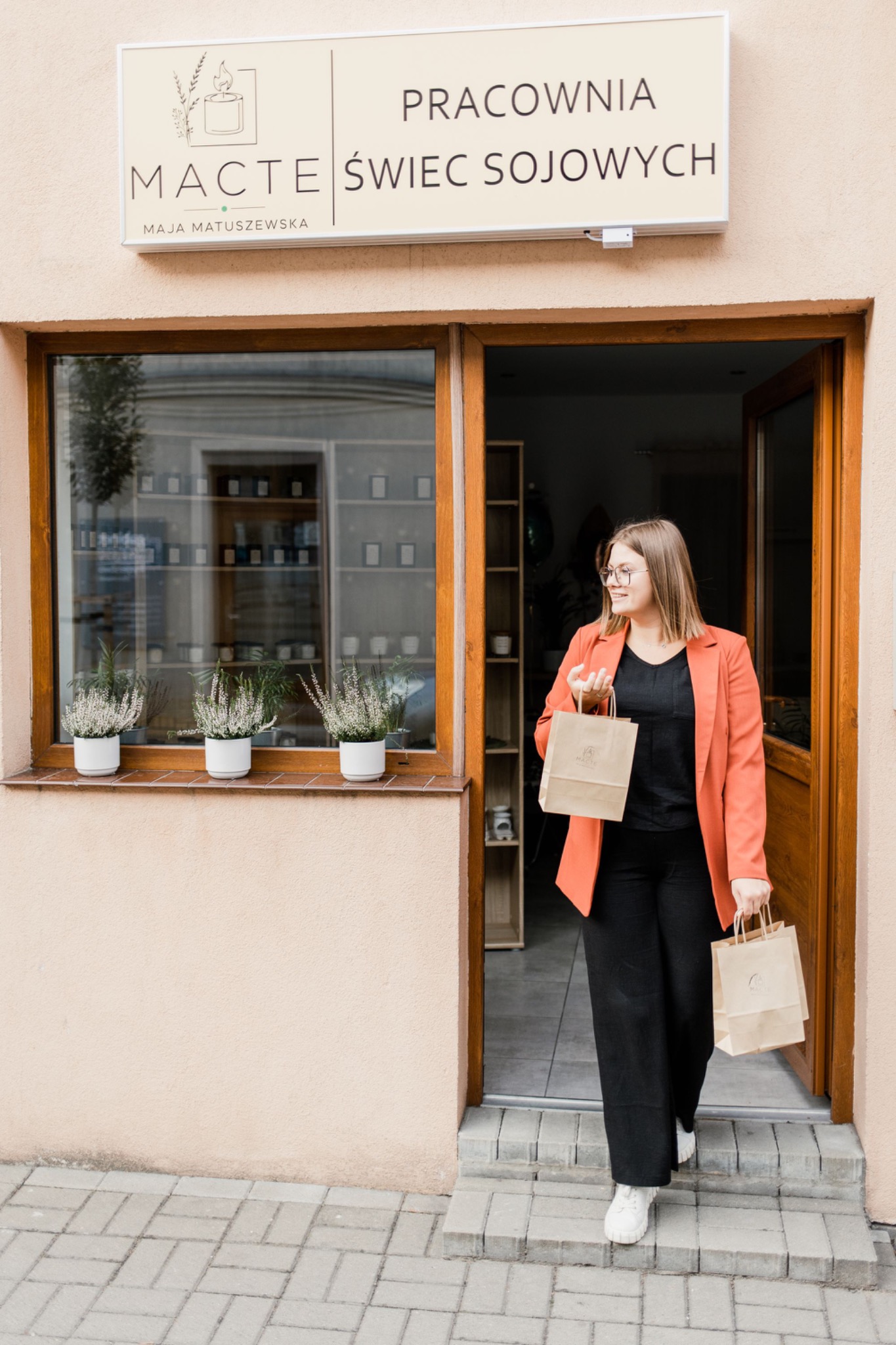Kobieta w koralowej marynarce wychodzi z pracowni świec sojowych, trzymając dwie papierowe torby z logo firmy.