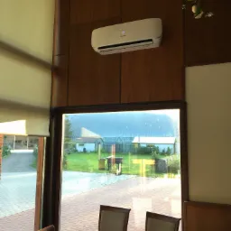 Biały klimatyzator Hisense zamontowany na drewnianej ścianie w jasnym pomieszczeniu z widokiem na ogród przez okno; na klimatyzatorze widoczny wyświetlacz z temperaturą 18 stopni.