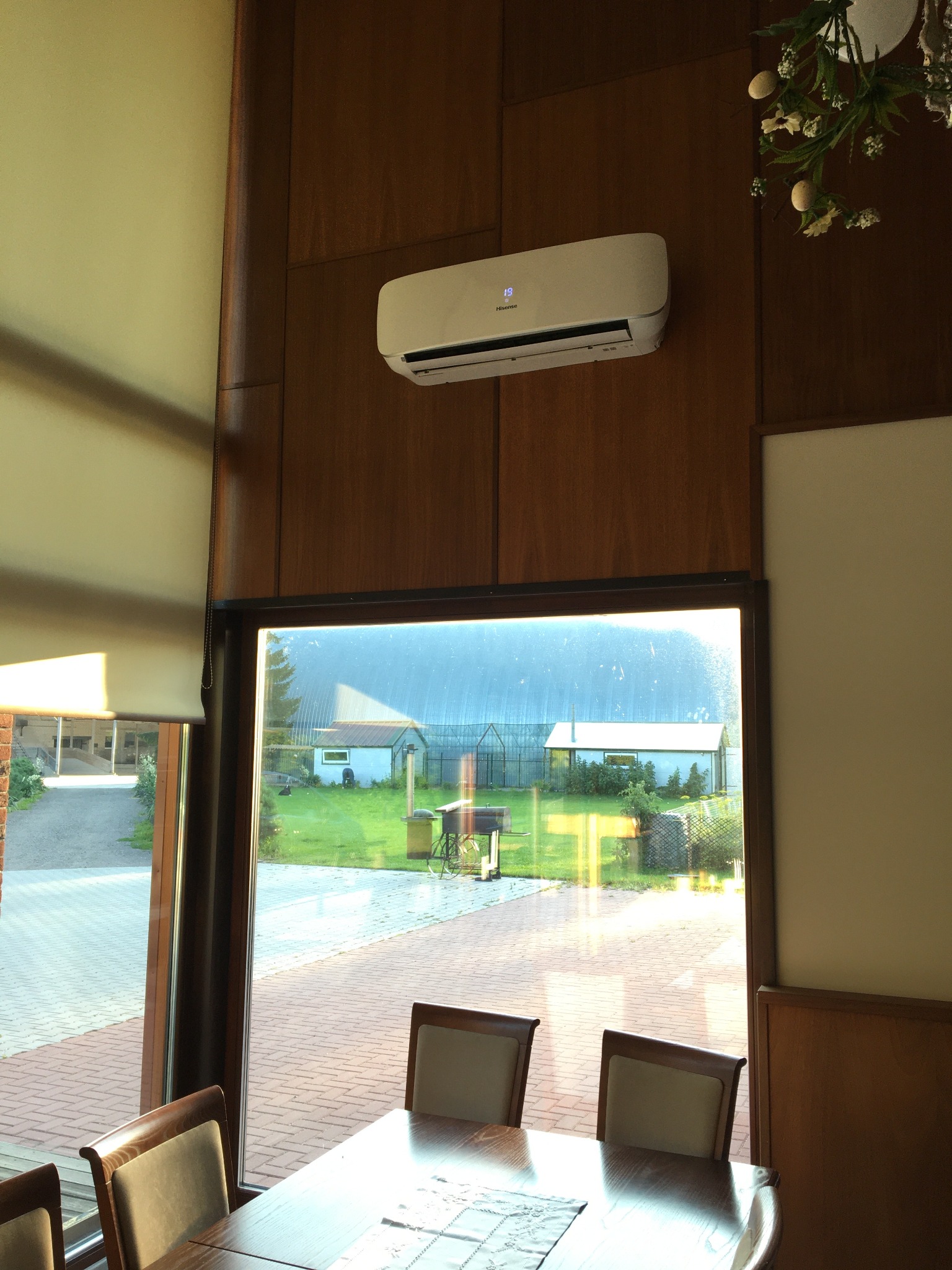 Biały klimatyzator Hisense zamontowany na drewnianej ścianie w jasnym pomieszczeniu z widokiem na ogród przez okno; na klimatyzatorze widoczny wyświetlacz z temperaturą 18 stopni.