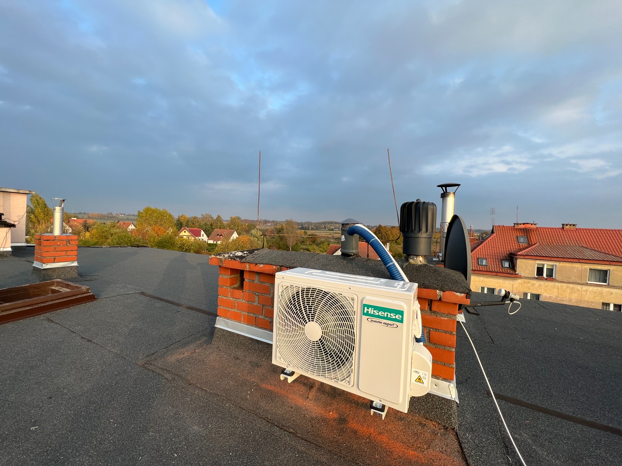 Jednostka zewnętrzna klimatyzacji Hisense zamontowana na dachu budynku, wsparta na metalowych wspornikach z pomarańczowymi śladami montażu, z widocznym niebieskim przewodem i kominem w tle, przy...