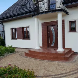 Biała elewacja domu jednorodzinnego z brązowymi oknami i drzwiami, kolumnami przy wejściu oraz brukowanym podjazdem z roślinnością.