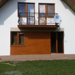 Ocieplony dom jednorodzinny z białymi ścianami, brązowymi oknami i balkonem z czarną balustradą, widoczny satelitarny talerz antenowy.