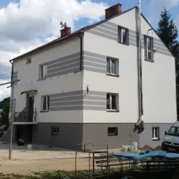 Dwukondygnacyjny dom po remoncie elewacji, z szarymi pasami na białej fasadzie, widoczne ślady prac wykończeniowych, w tle błękitne niebo z chmurami.