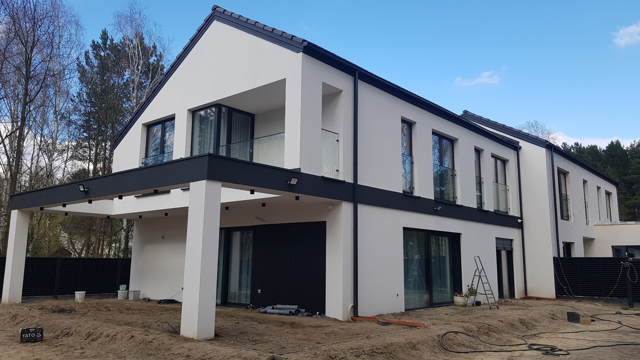 Nowoczesny dom jednorodzinny z białą elewacją, czarnymi detalami architektonicznymi i szklanymi balustradami balkonów, widoczne ślady prac wykończeniowych w postaci rur i drabiny na posesji.
