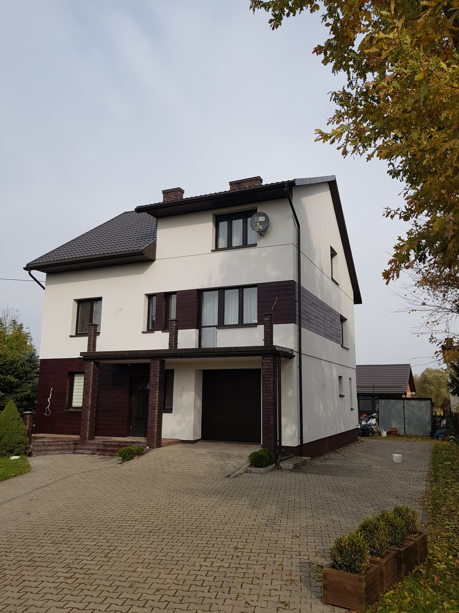 Dwukondygnacyjny dom z białymi ścianami i brązowymi elementami dekoracyjnymi, brązowy dach, kostka brukowa na podjeździe, antena satelitarna na elewacji.