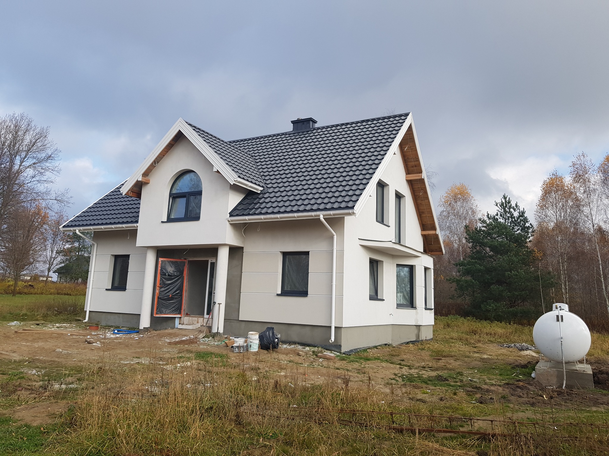 Nowo otynkowany, dwukondygnacyjny dom z ciemną dachówką i białymi rynnami, z widocznymi śladami prac budowlanych wokół, w tym wiadra i folia zabezpieczająca wejście.