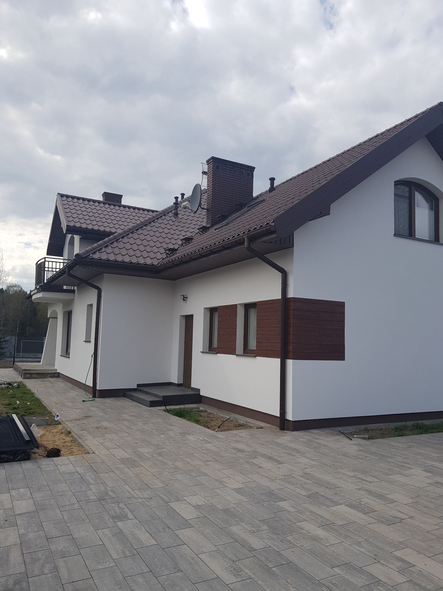 Nowoczesny dom jednorodzinny z białą elewacją, brązowym dachem i drewnianymi akcentami, widok z kostki brukowej na tle pochmurnego nieba.