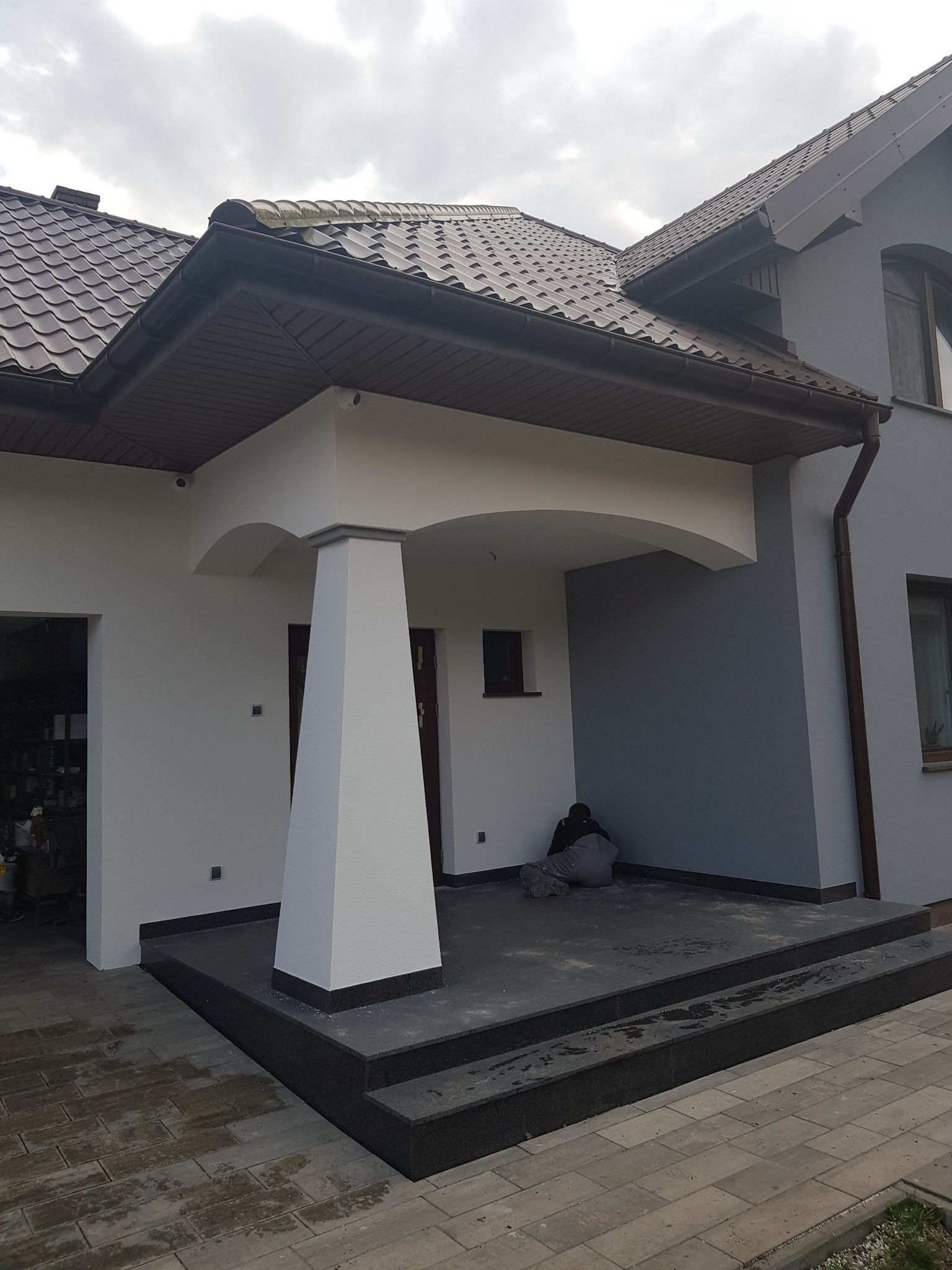 Dom jednorodzinny po odświeżeniu elewacji, z widocznym fragmentem tarasu, gdzie pracownik w roboczym ubraniu siedzi na ziemi, prawdopodobnie w trakcie sprzątania po pracach.