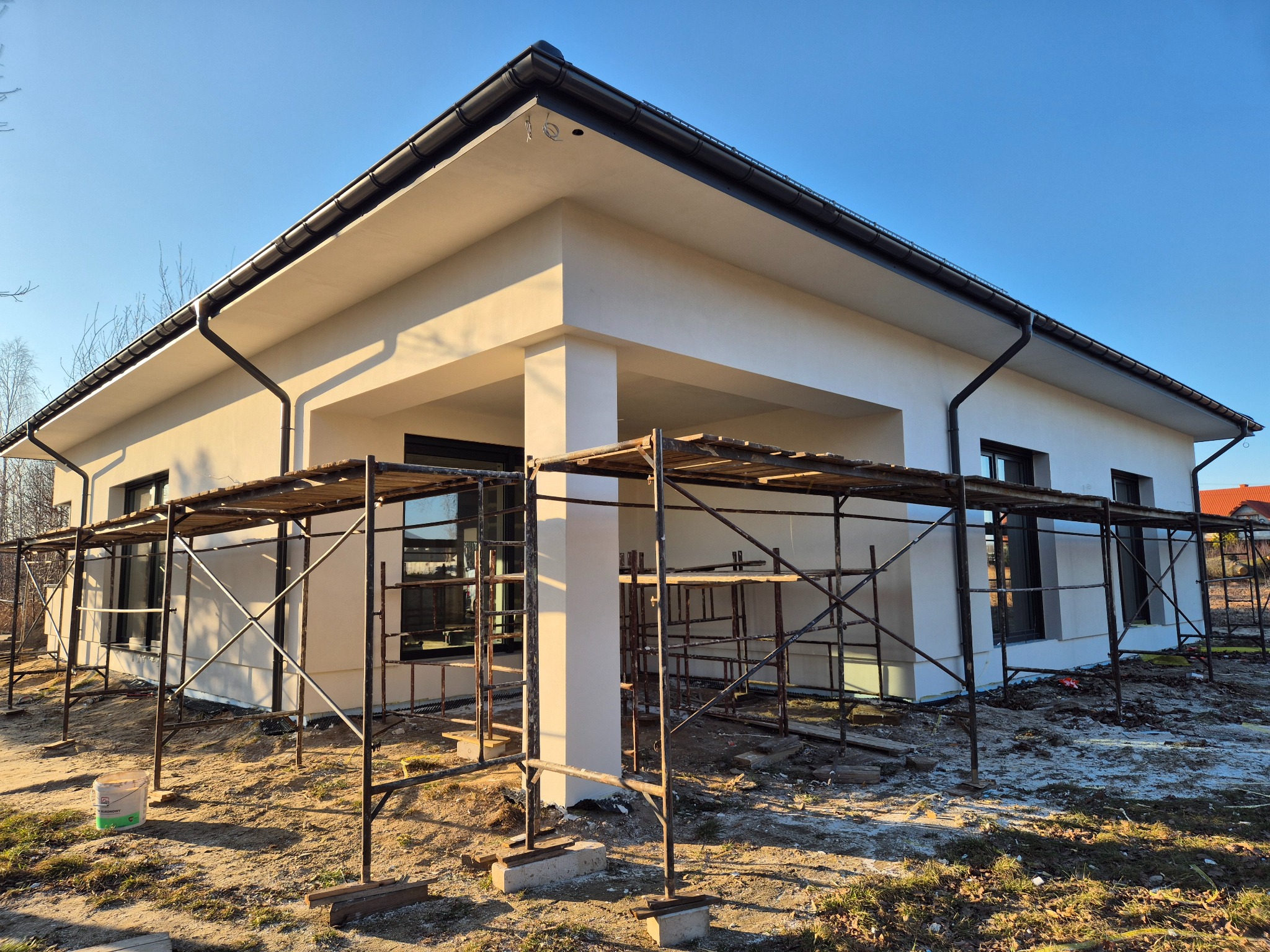 Nowoczesny dom jednorodzinny z jasną elewacją, częściowo osłonięty rusztowaniami, widoczne czarne rynny i okna, teren wokół domu w trakcie prac wykończeniowych.